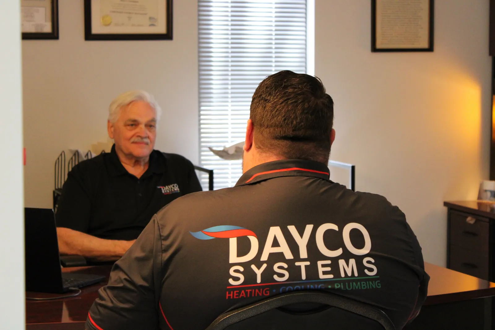 Technician with Dayco Systems logo on shirt consulting a senior man in an office setting with certificates on wall.