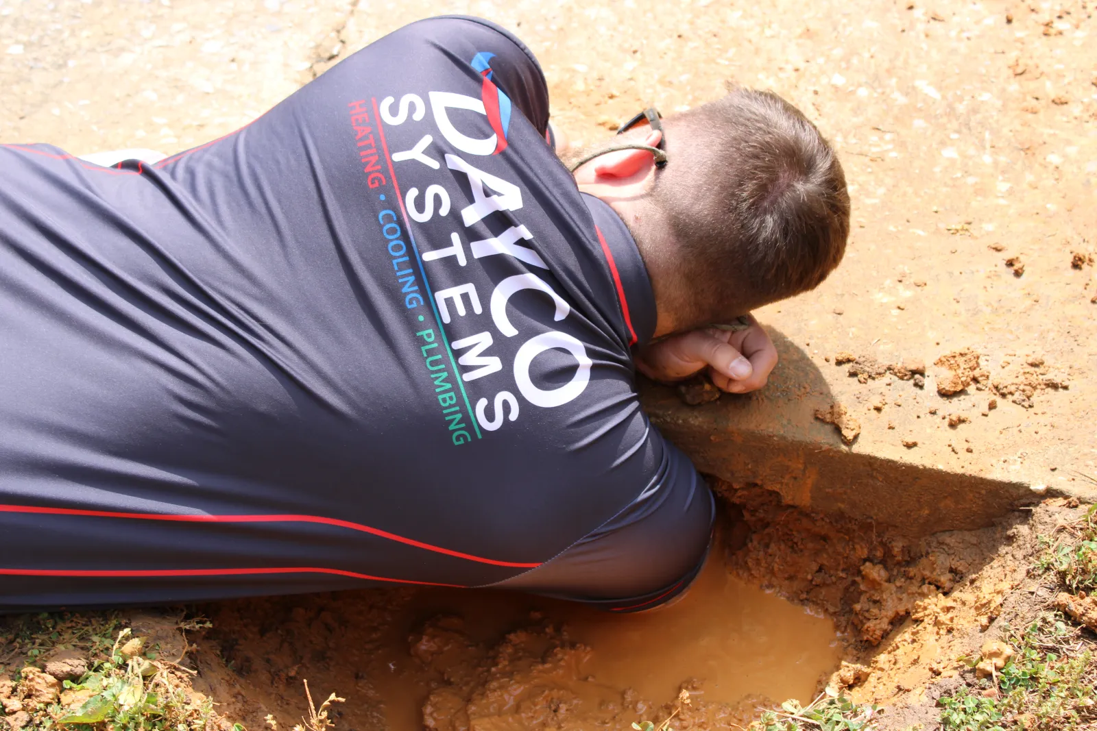 Technician from Dayco Systems inspecting a muddy outdoor plumbing or drainage site laying on the ground.