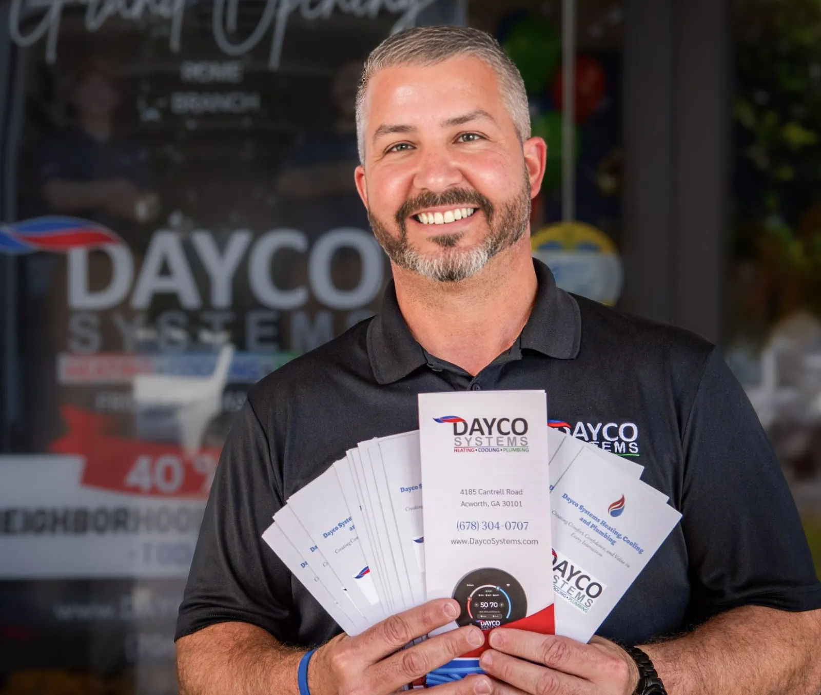 Smiling man in black Dayco Systems shirt holding fan of company brochures outside business entrance.