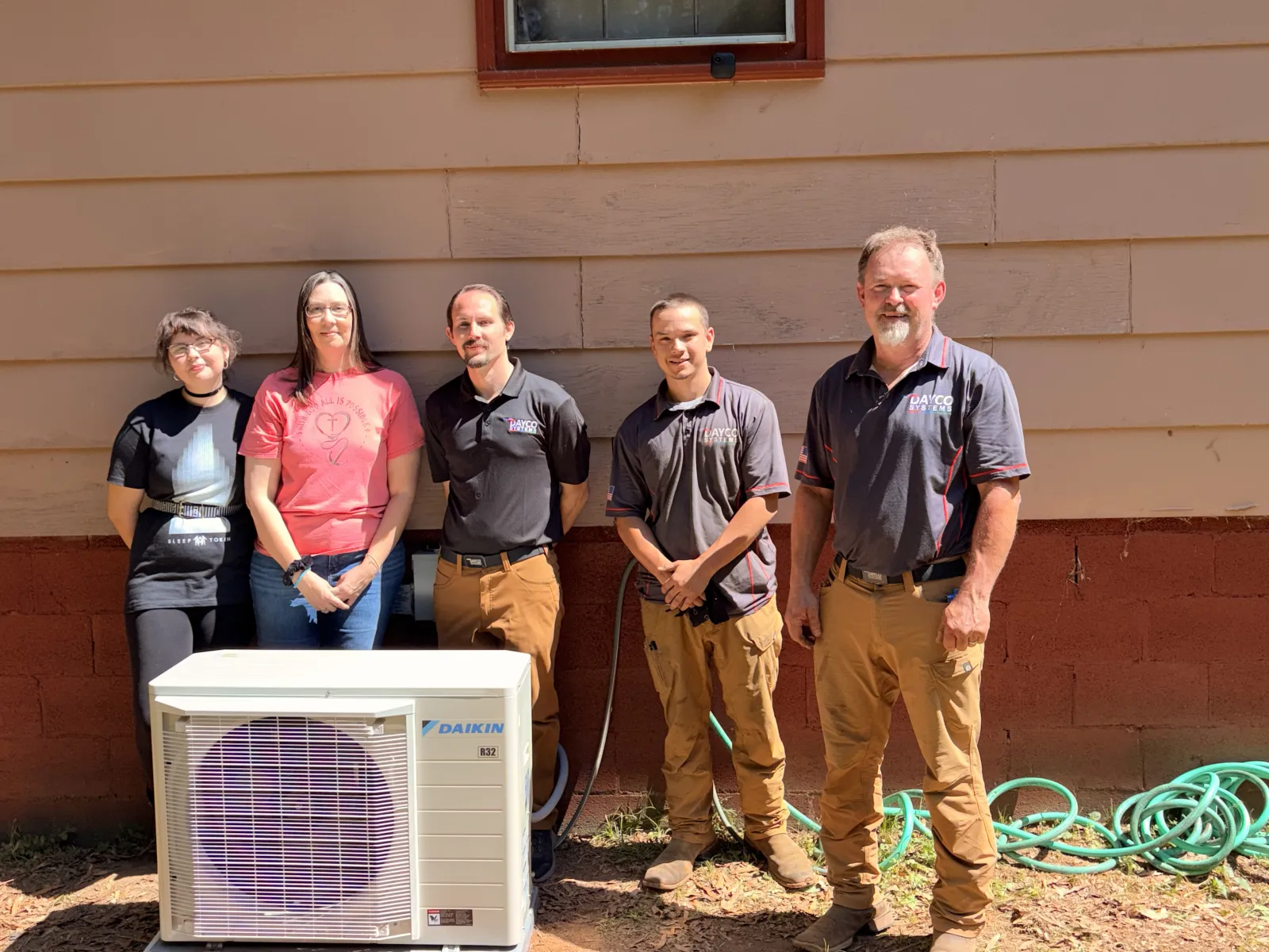 Five people standing outdoors behind a Daikin air conditioning unit next to a red and beige building wall.