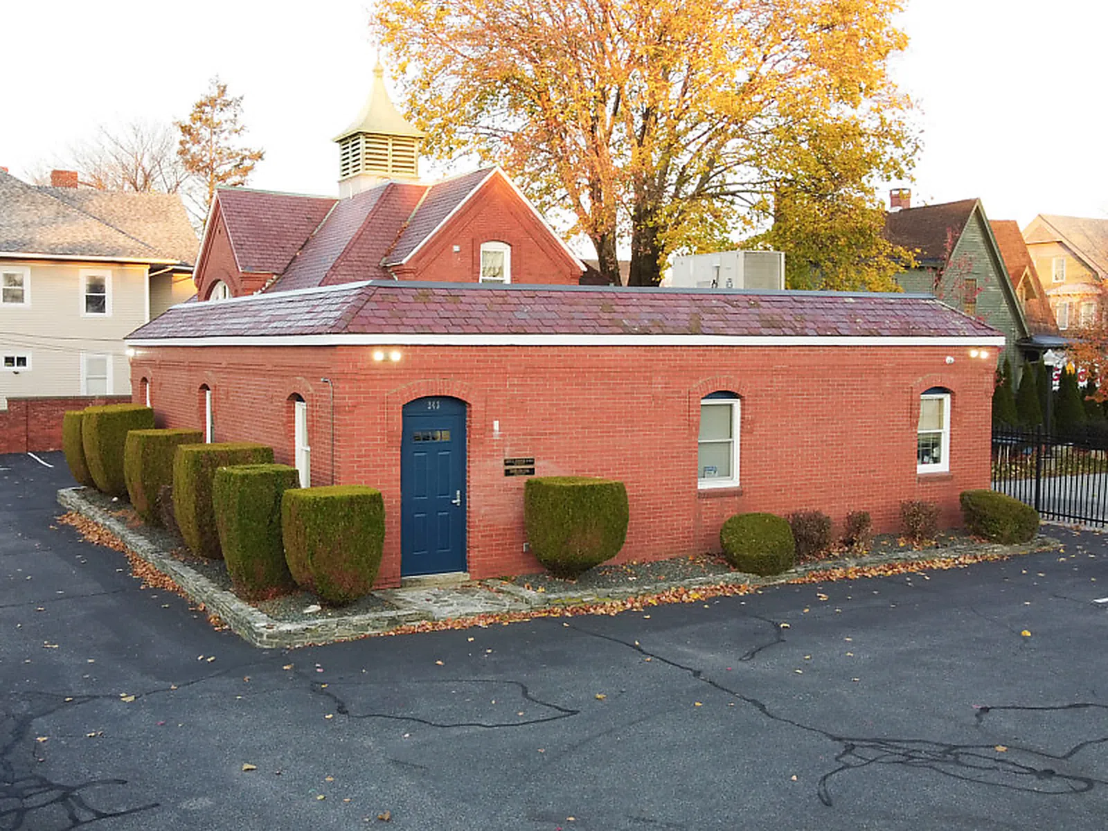 Small brick building with blue door, trimmed bushes, and large autumn tree behind in suburban area.