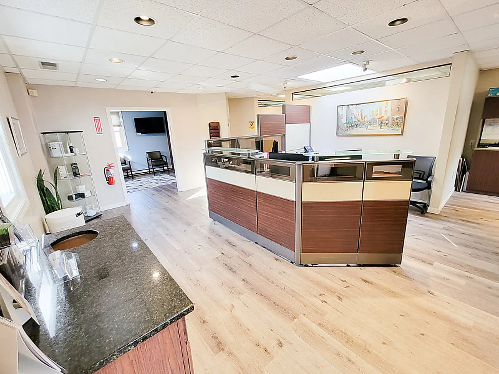 Bright modern office reception area with wooden flooring, glass panel desks, and seating in adjacent room.