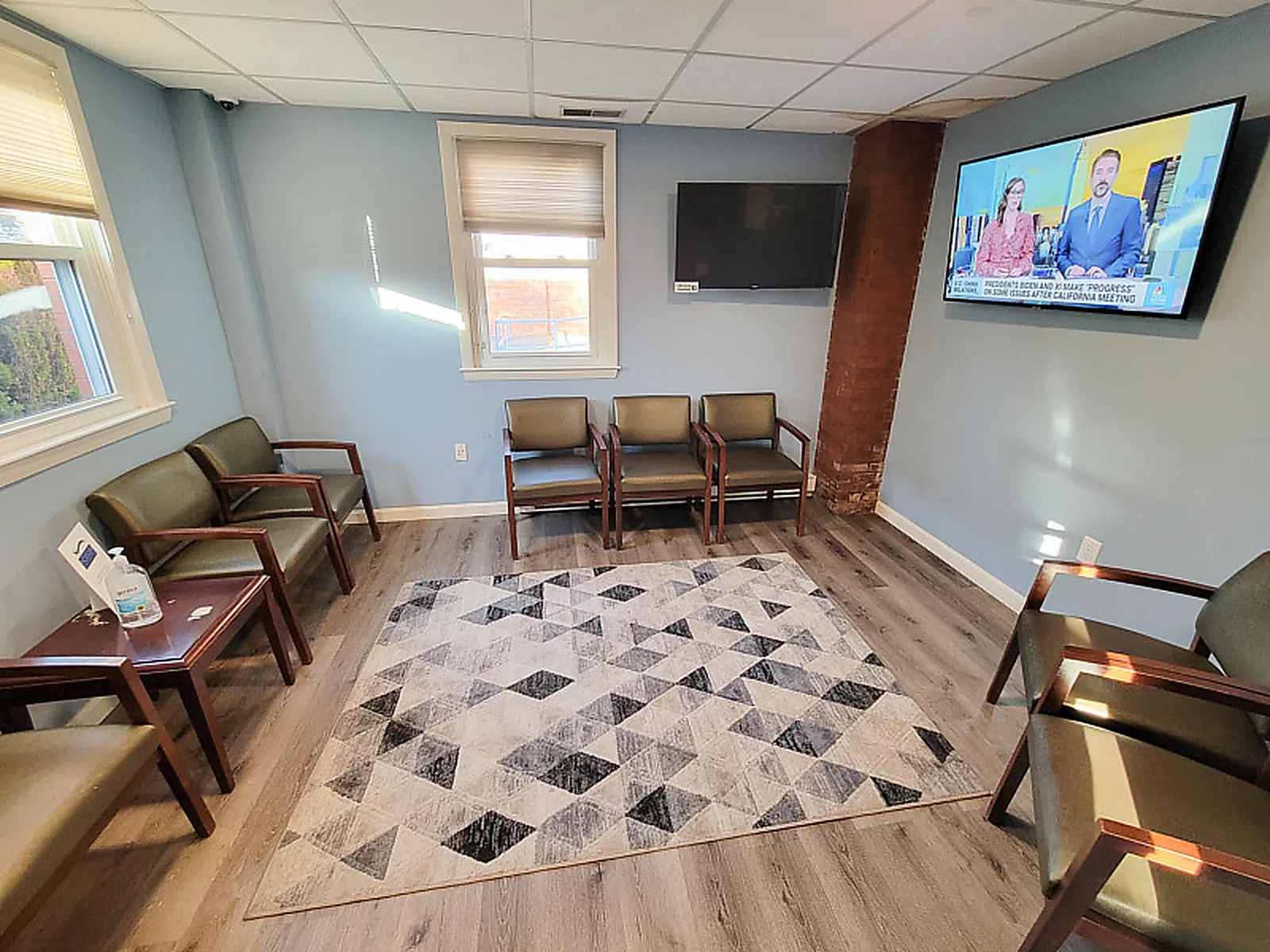 Bright waiting room with chairs, geometric rug, two windows, two TVs, and hardwood flooring under natural light