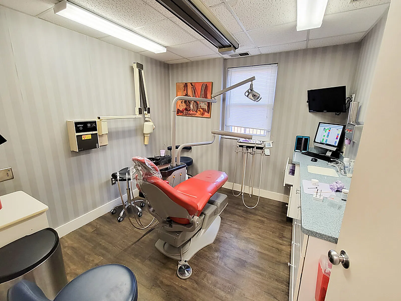 Modern dental exam room with red patient chair, dental tools, computer, and striped wallpaper.
