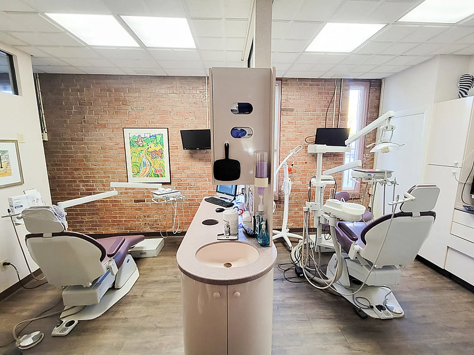 Modern dental office with two dental chairs, equipment, sink, and brick accent walls under bright lighting.