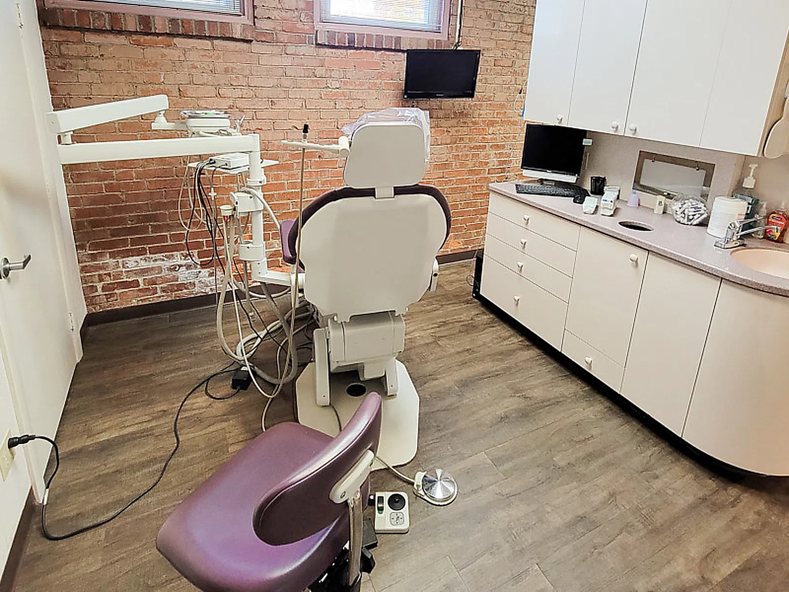 Modern dental treatment room with dental chair, equipment, brick wall, and countertop with supplies.