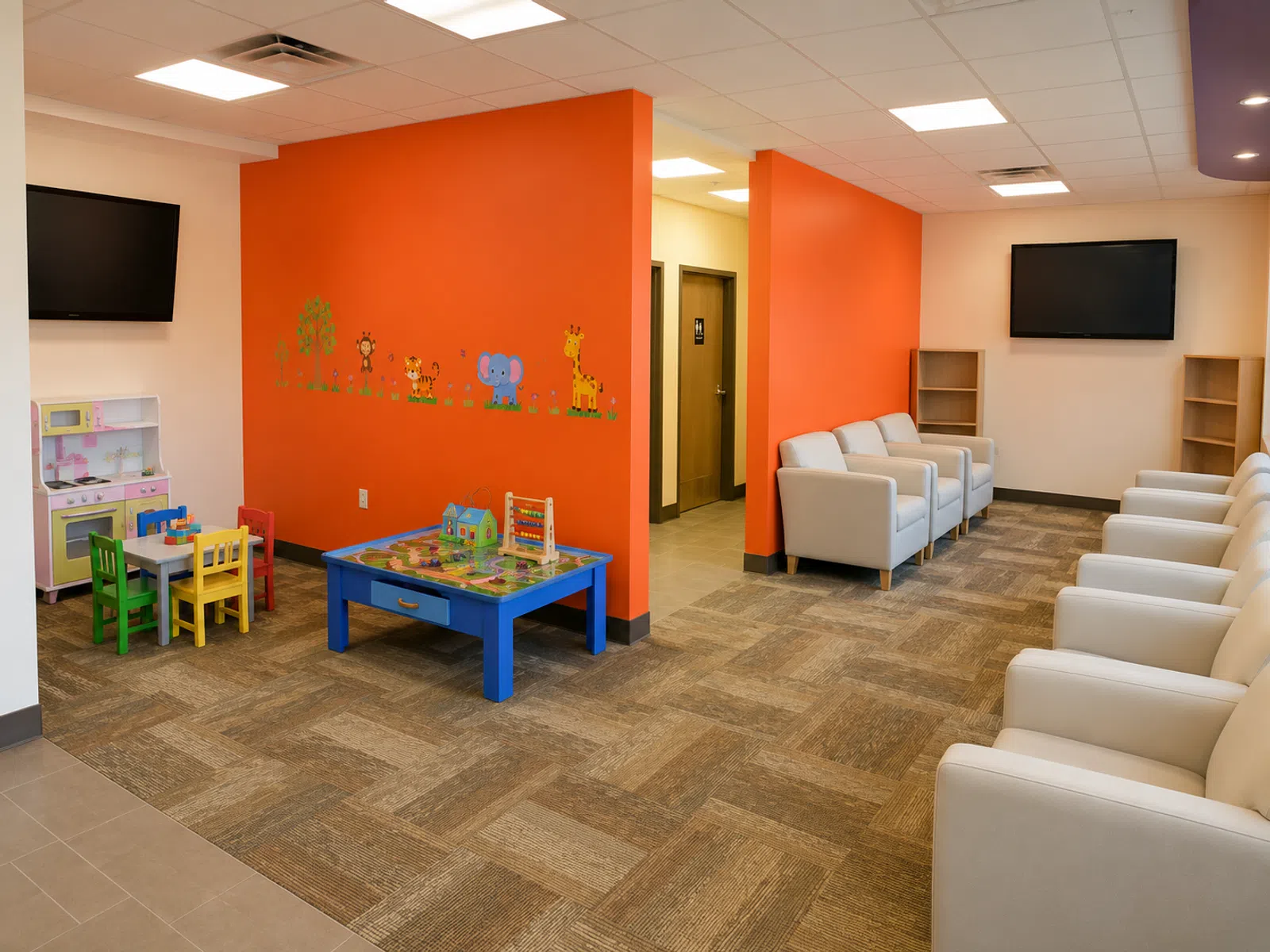Bright waiting room with white chairs, orange walls, a TV, and a colorful children's play area with toys and chairs.
