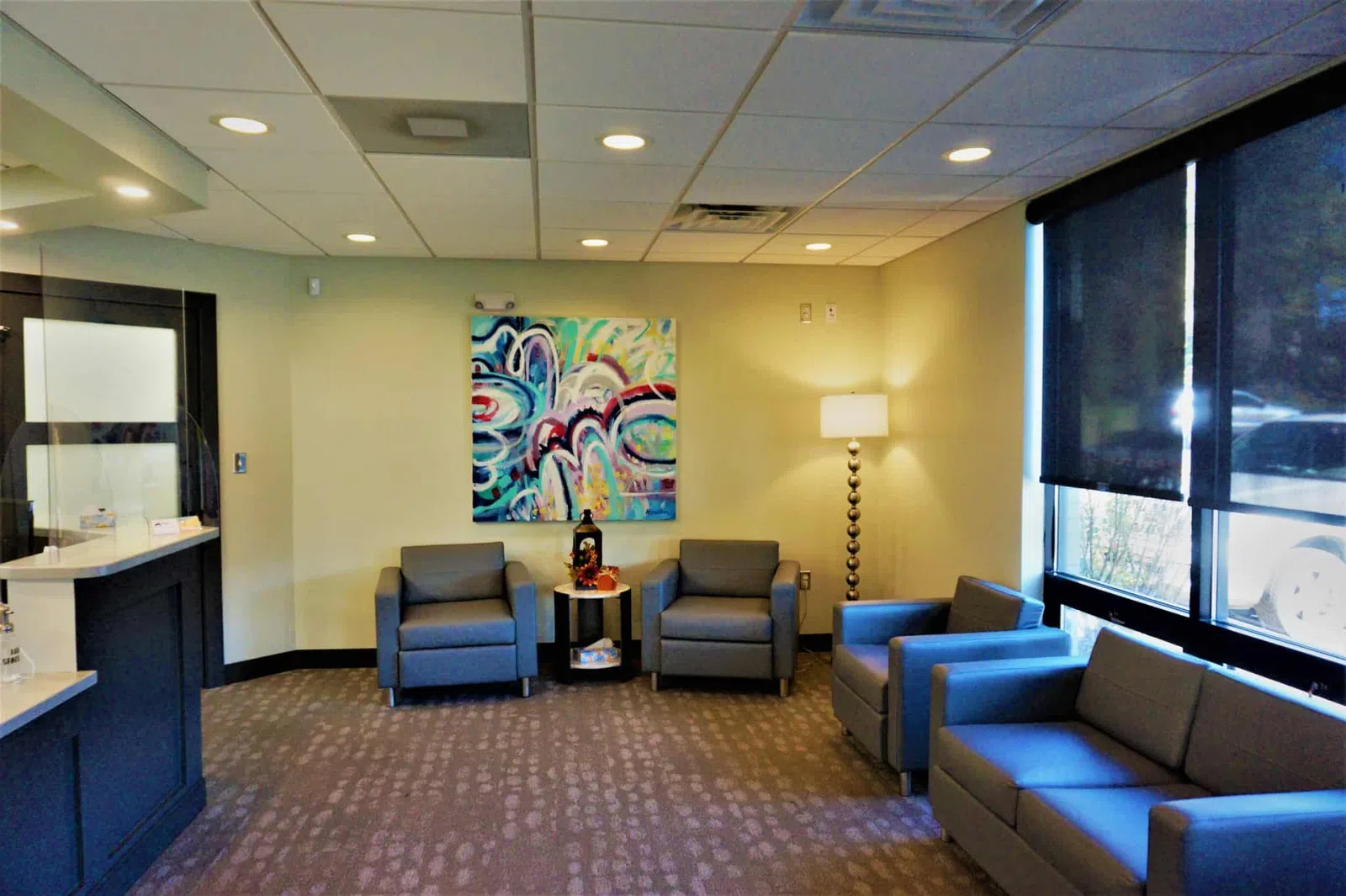 Modern waiting room with gray chairs, colorful abstract painting, floor lamp, and large windows with dark blinds.