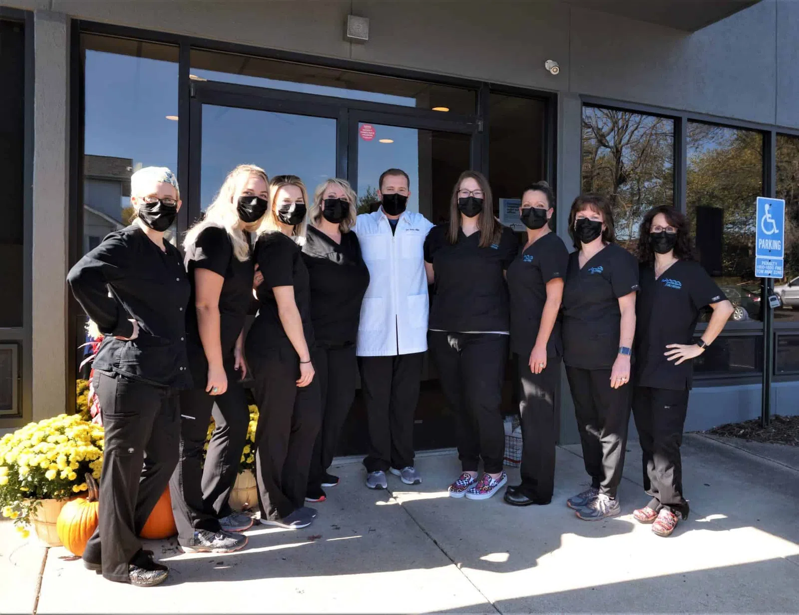 Group of healthcare workers wearing black scrubs and masks standing outside a clinic entrance with autumn decorations.
