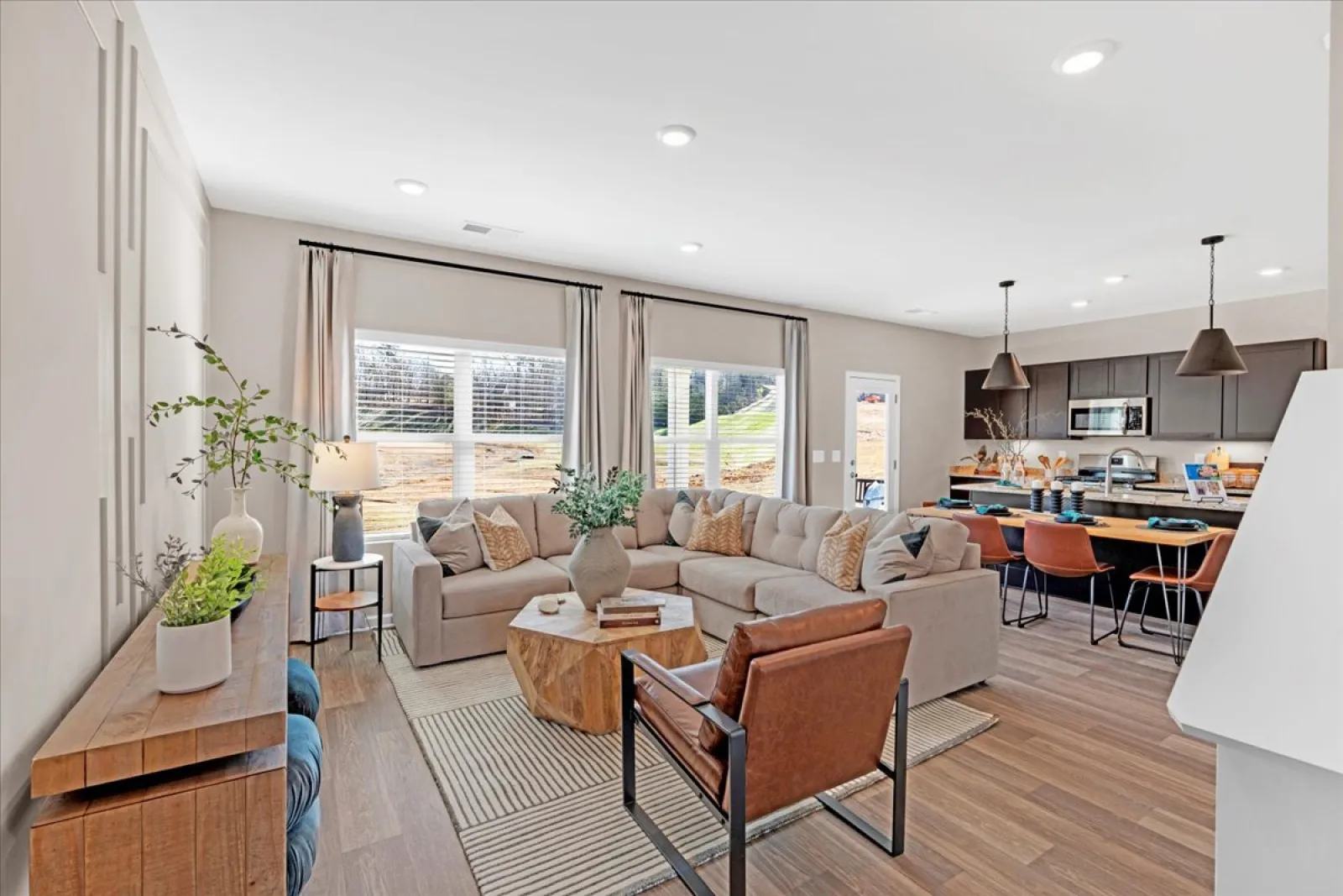 Modern living room with beige sectional sofa, wood accents, and large windows overlooking green yard.