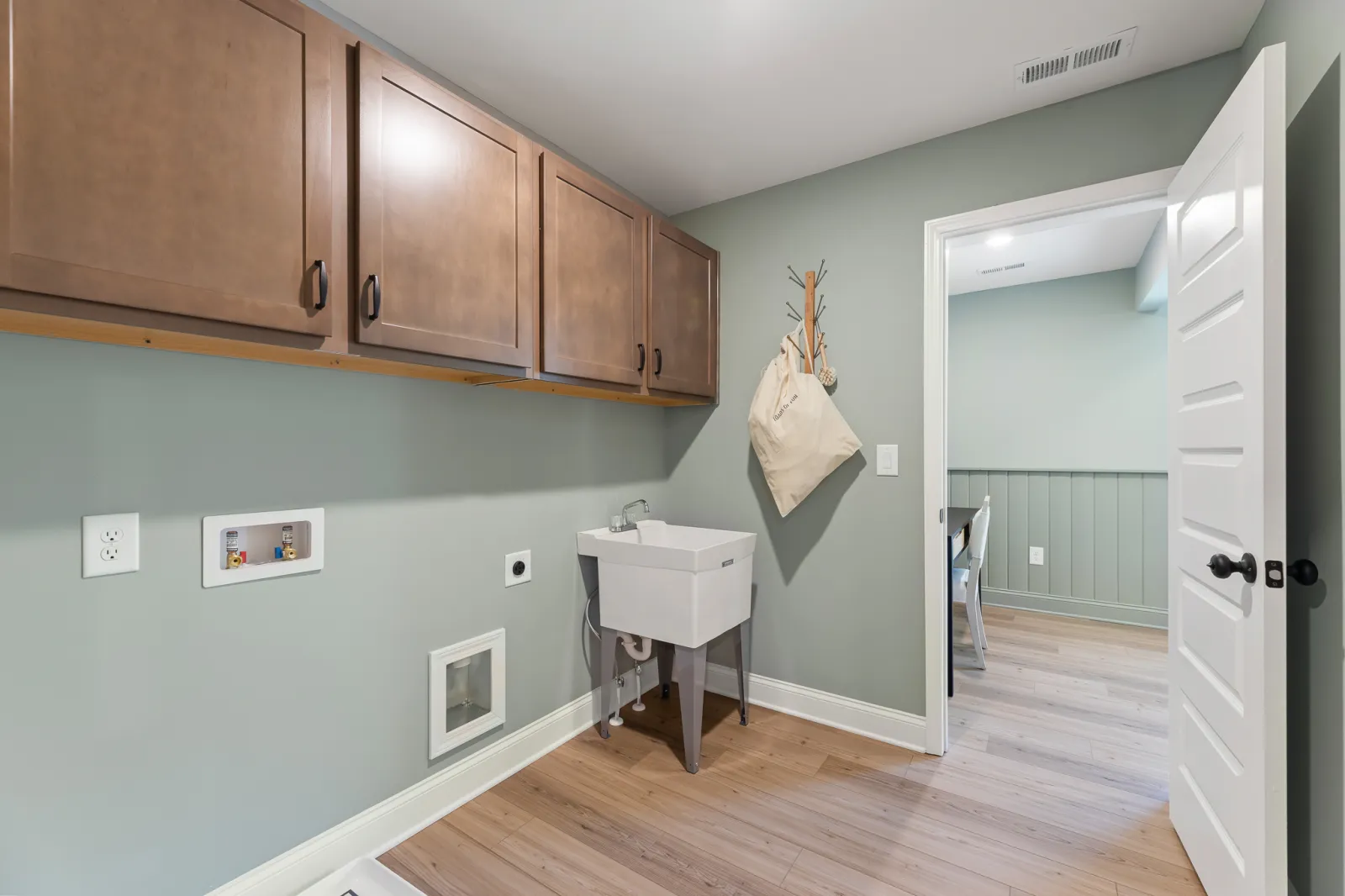 Modern laundry room with wooden cabinets, utility sink, light green walls, and hardwood flooring next to an open door.