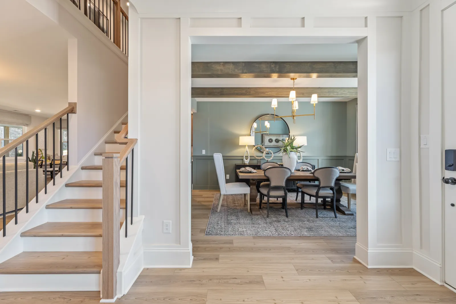View of modern dining room with wooden beams, gray walls, chandelier, and light wood staircase and flooring.
