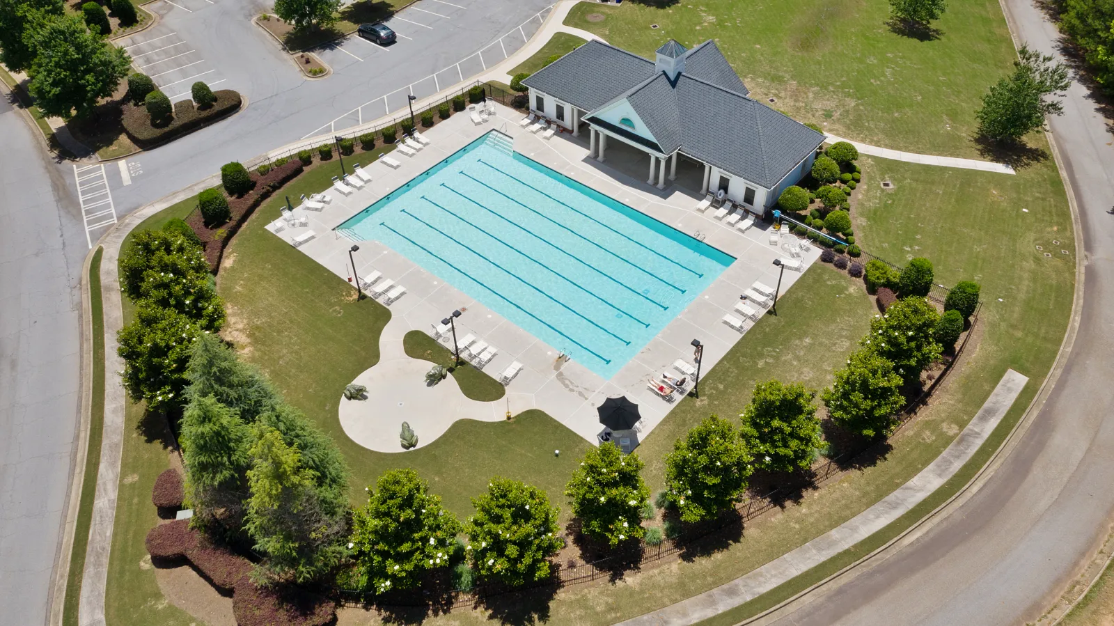 Aerial view of a rectangular outdoor swimming pool with lounge chairs and a nearby building surrounded by trees.