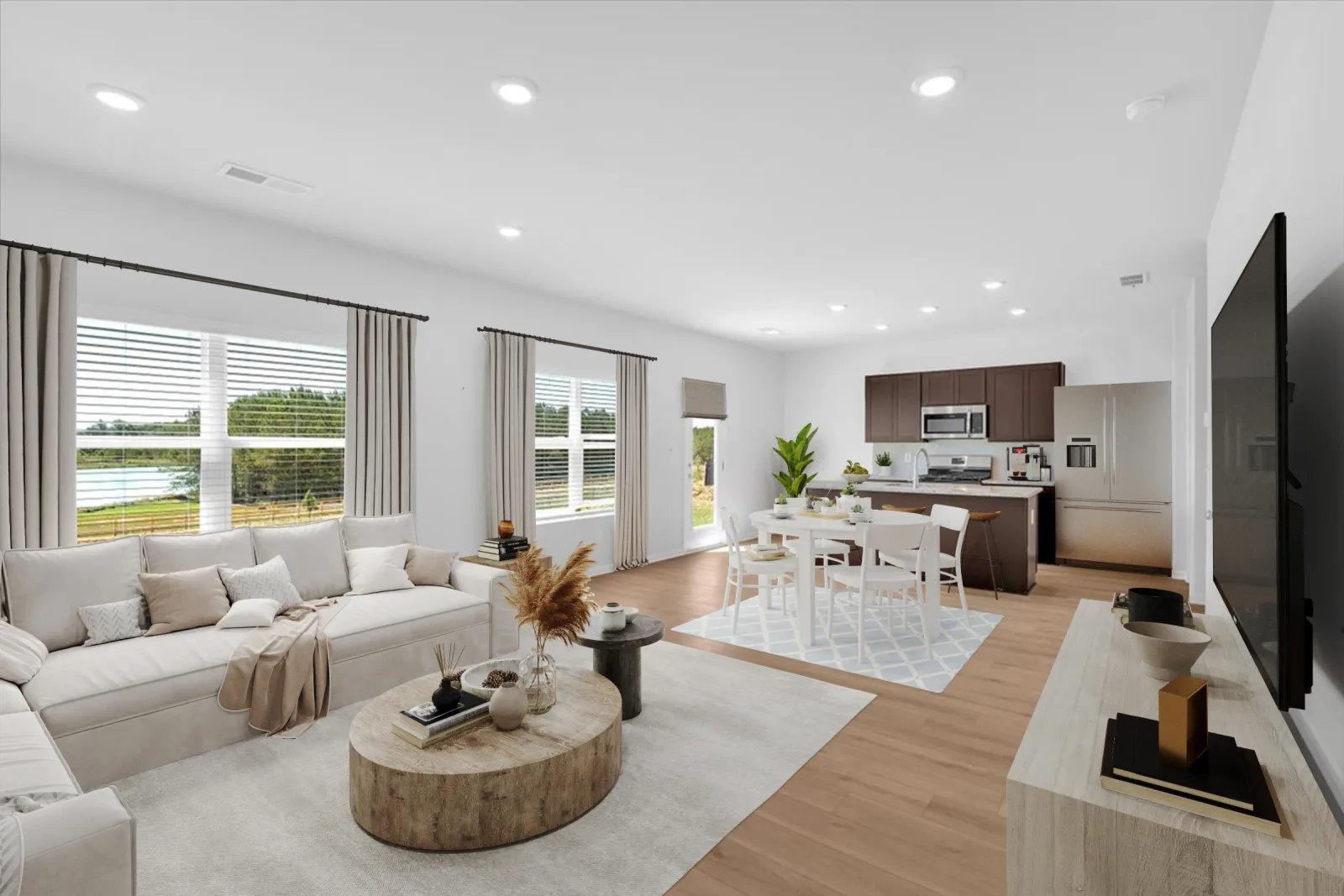 Bright open-concept living room and kitchen with white furniture, wooden floors, and large windows showcasing outdoor greenery.