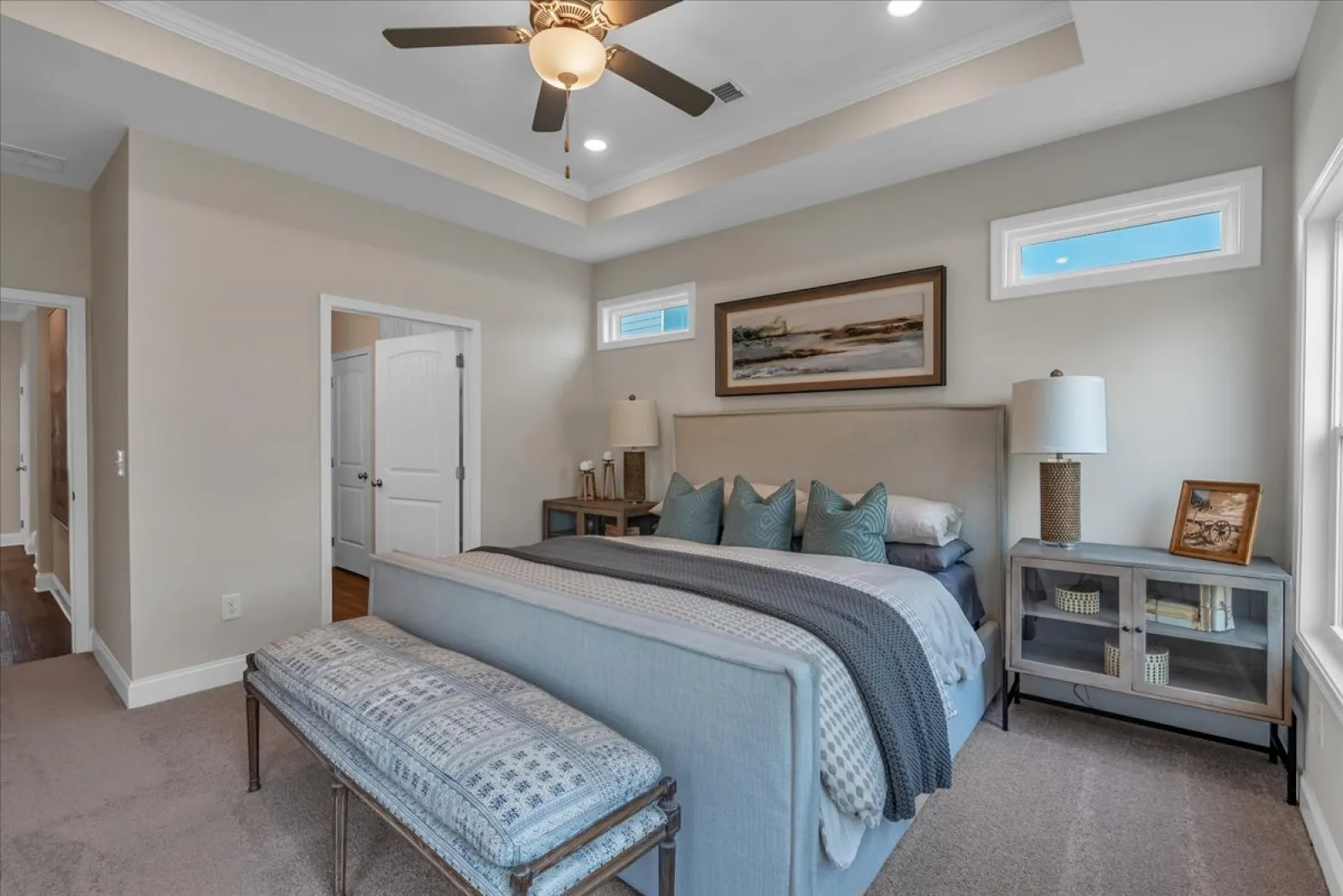 Modern bedroom with a king-size bed, blue and gray bedding, bench, lamps, and ceiling fan under tray ceiling.
