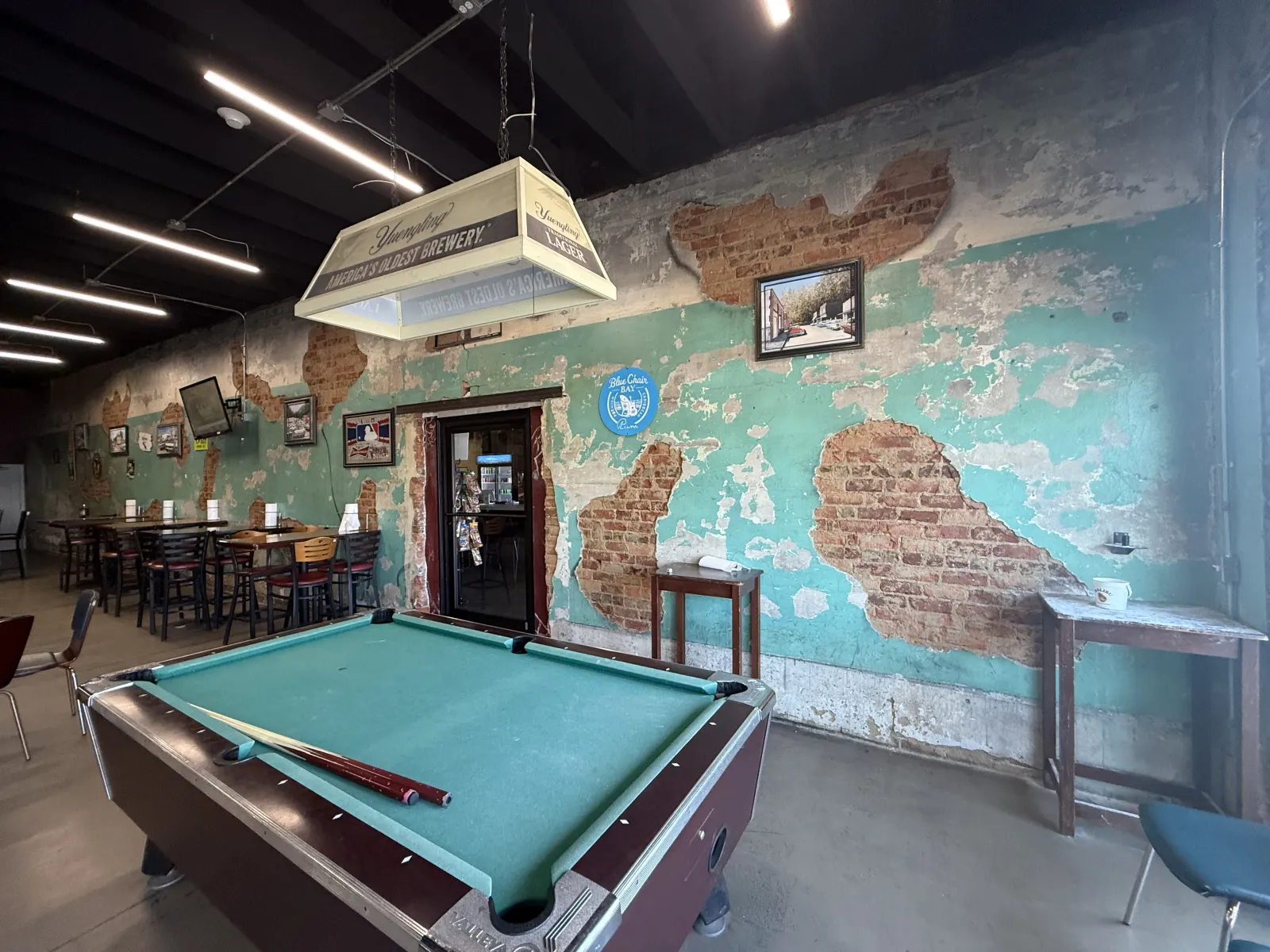 Pool table in a rustic bar with exposed brick and peeling turquoise paint walls, wooden furniture, and vintage decor.
