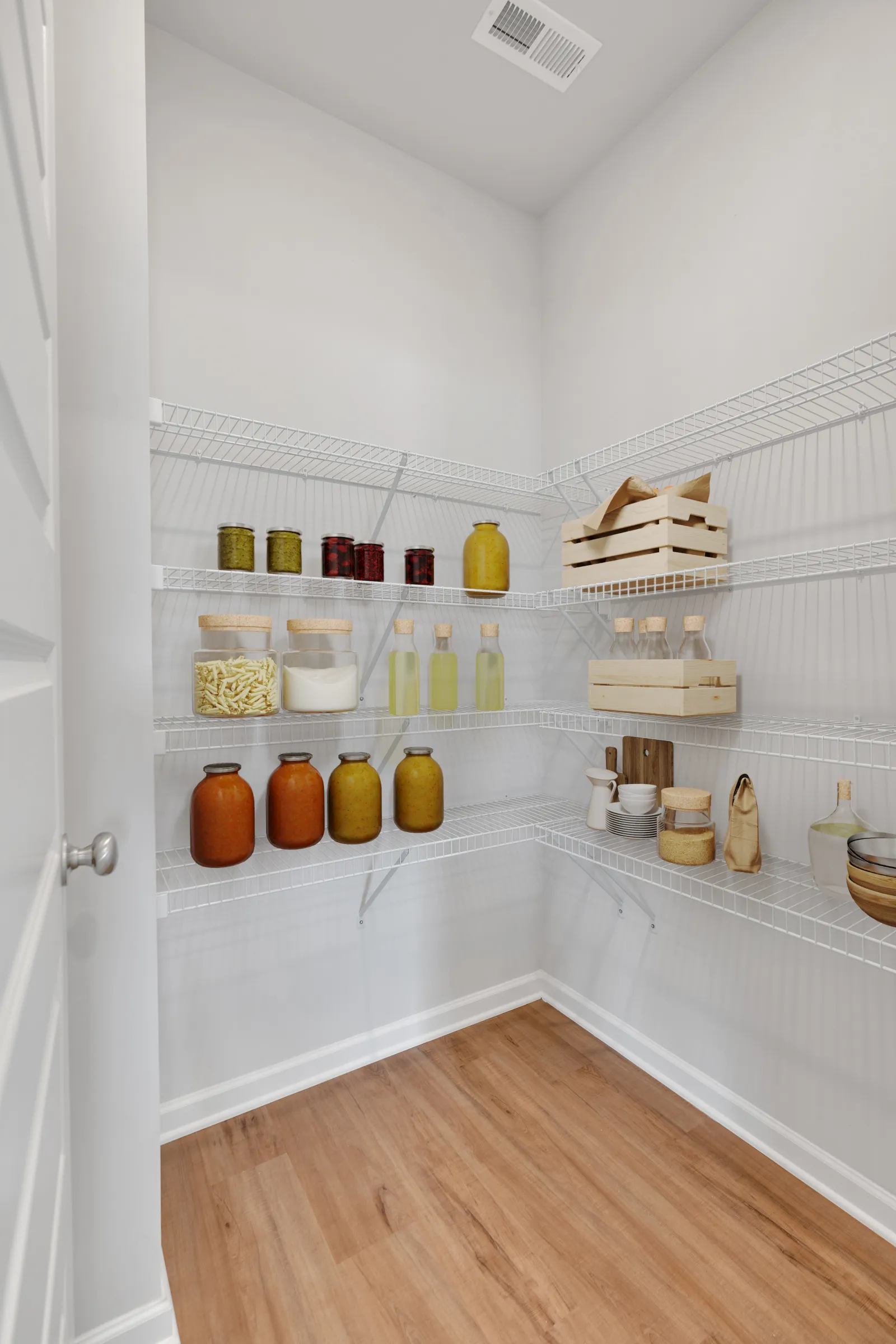 Organized pantry with white wire shelves holding jars, bottles, wooden crates, and kitchenware on wood floor.