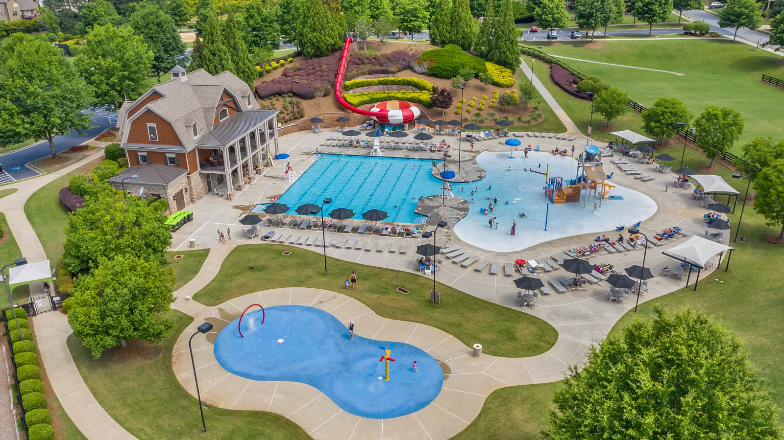 Aerial view of a public pool complex with swimming pool, water play area, slide, and splash pad, surrounded by green parkland.