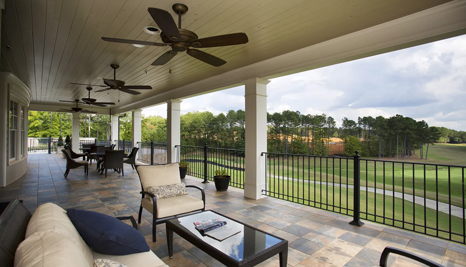Spacious covered patio with ceiling fans overlooking a lush golf course and surrounded by trees under a cloudy sky