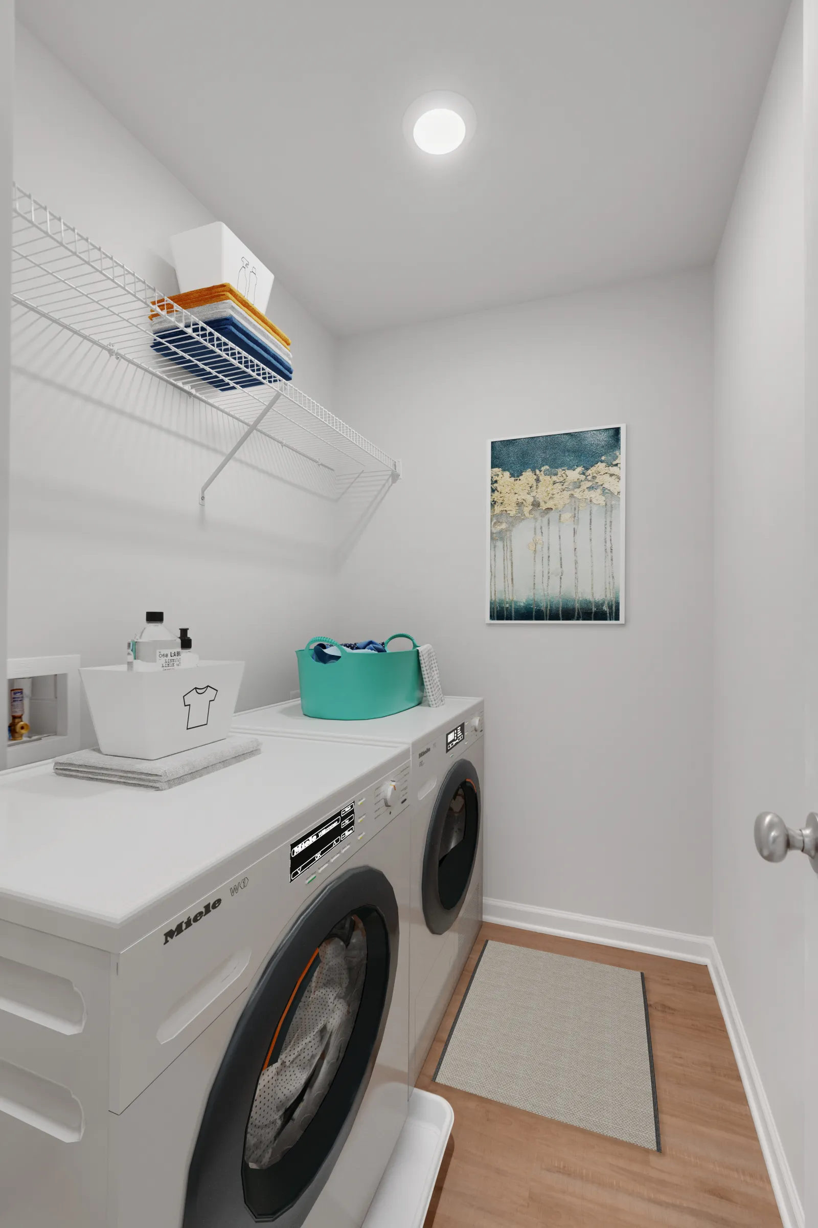 Compact laundry room with front-loading washer and dryer, white shelves, storage bins, and wall art.