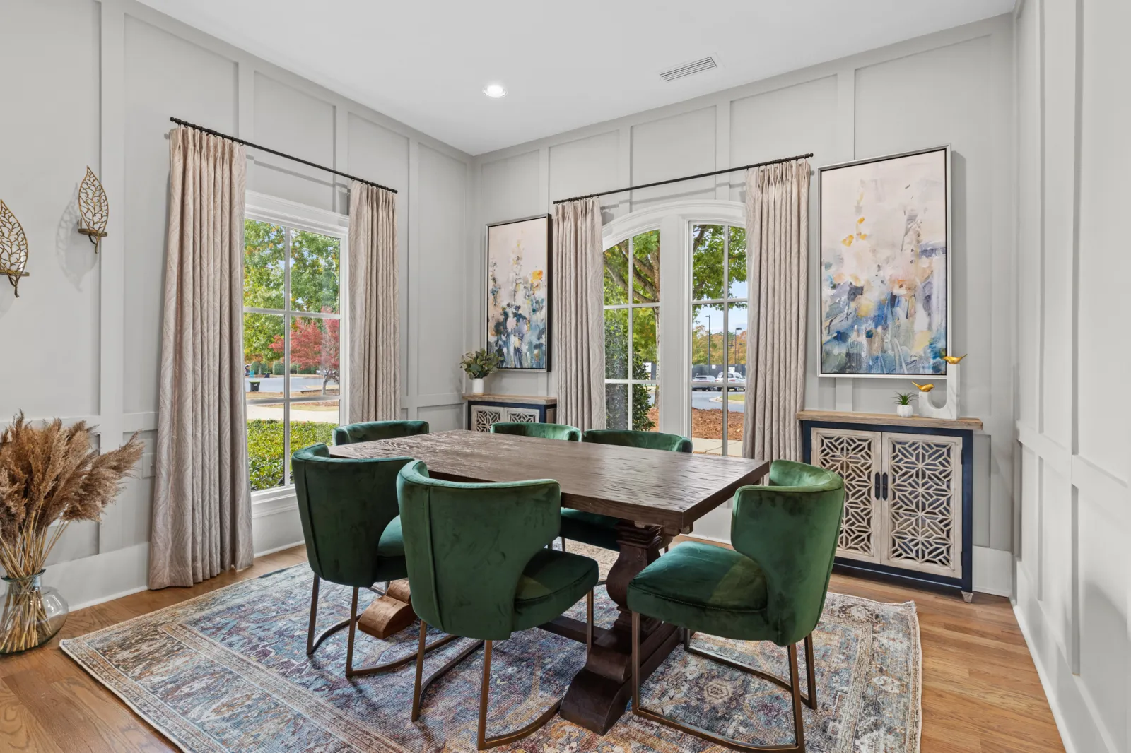 Bright dining room with wooden table, green velvet chairs, patterned rug, large windows, and abstract wall art.