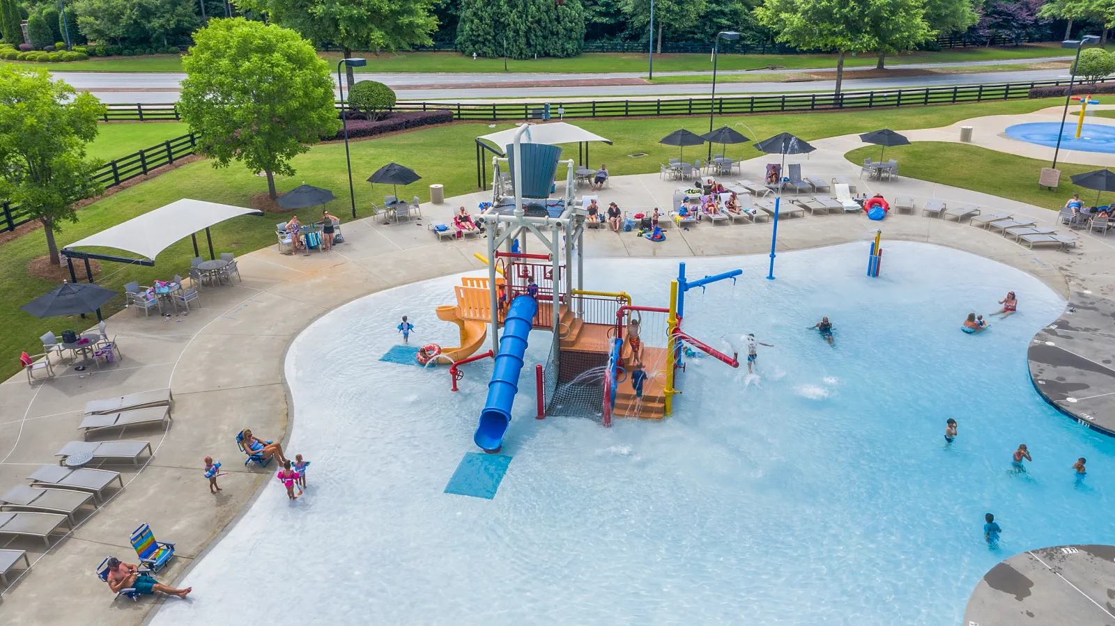 Outdoor water park with a splash pad, slides, and people enjoying in shallow pool under umbrellas.