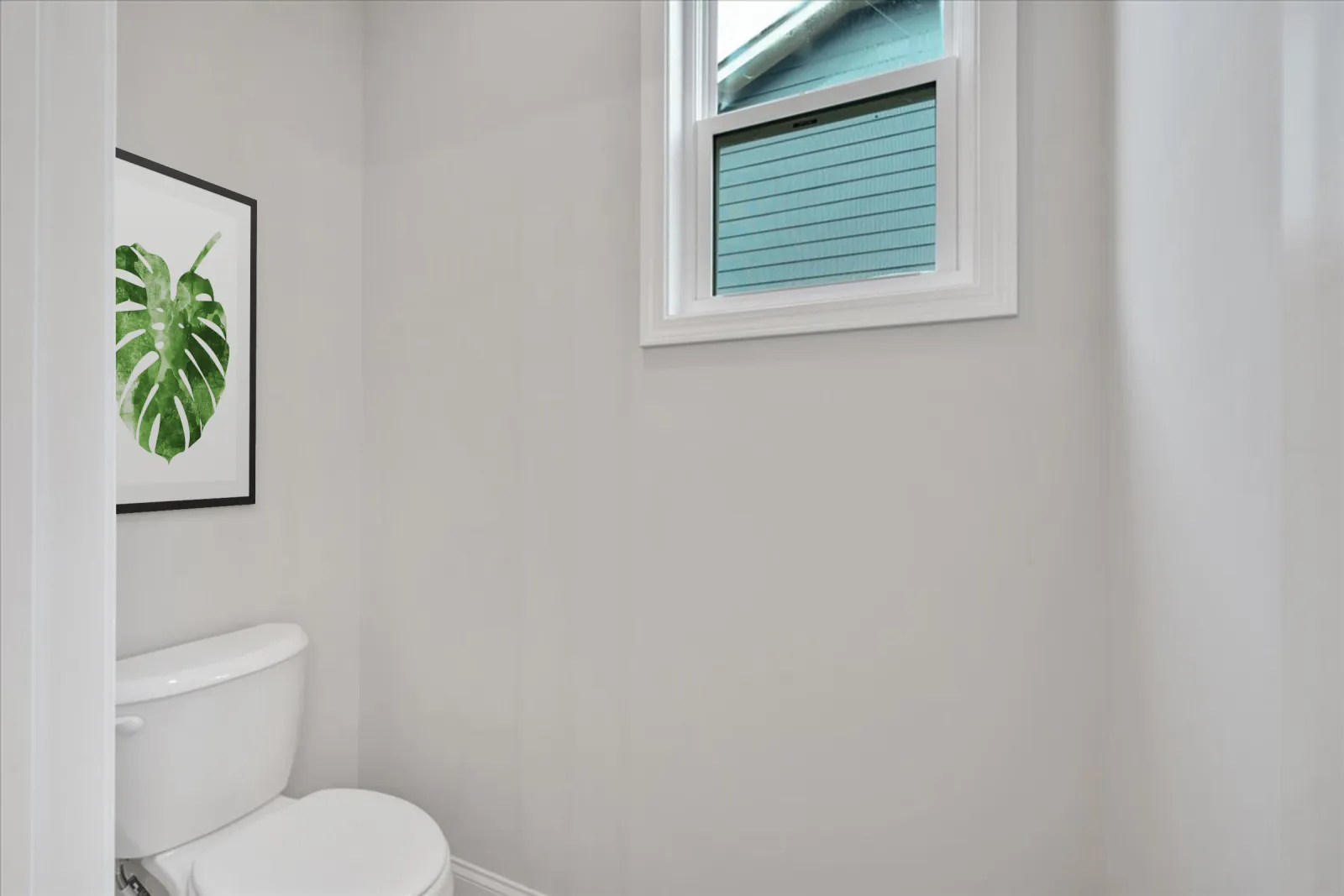 Minimalist bathroom with white toilet, gray walls, a window, and framed green leaf artwork.