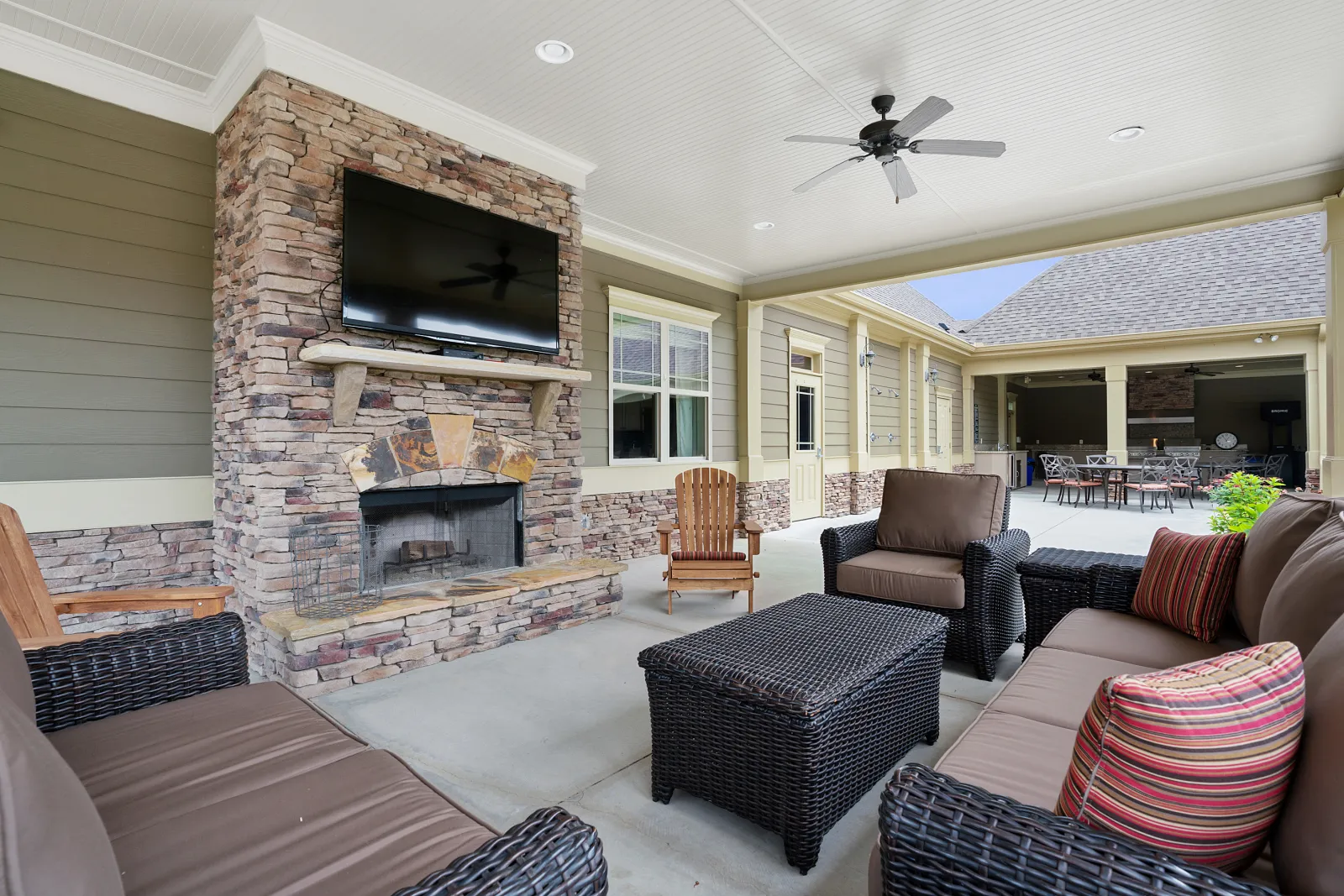 Cozy outdoor patio with stone fireplace, mounted TV, rattan furniture, and ceiling fan under covered porch.
