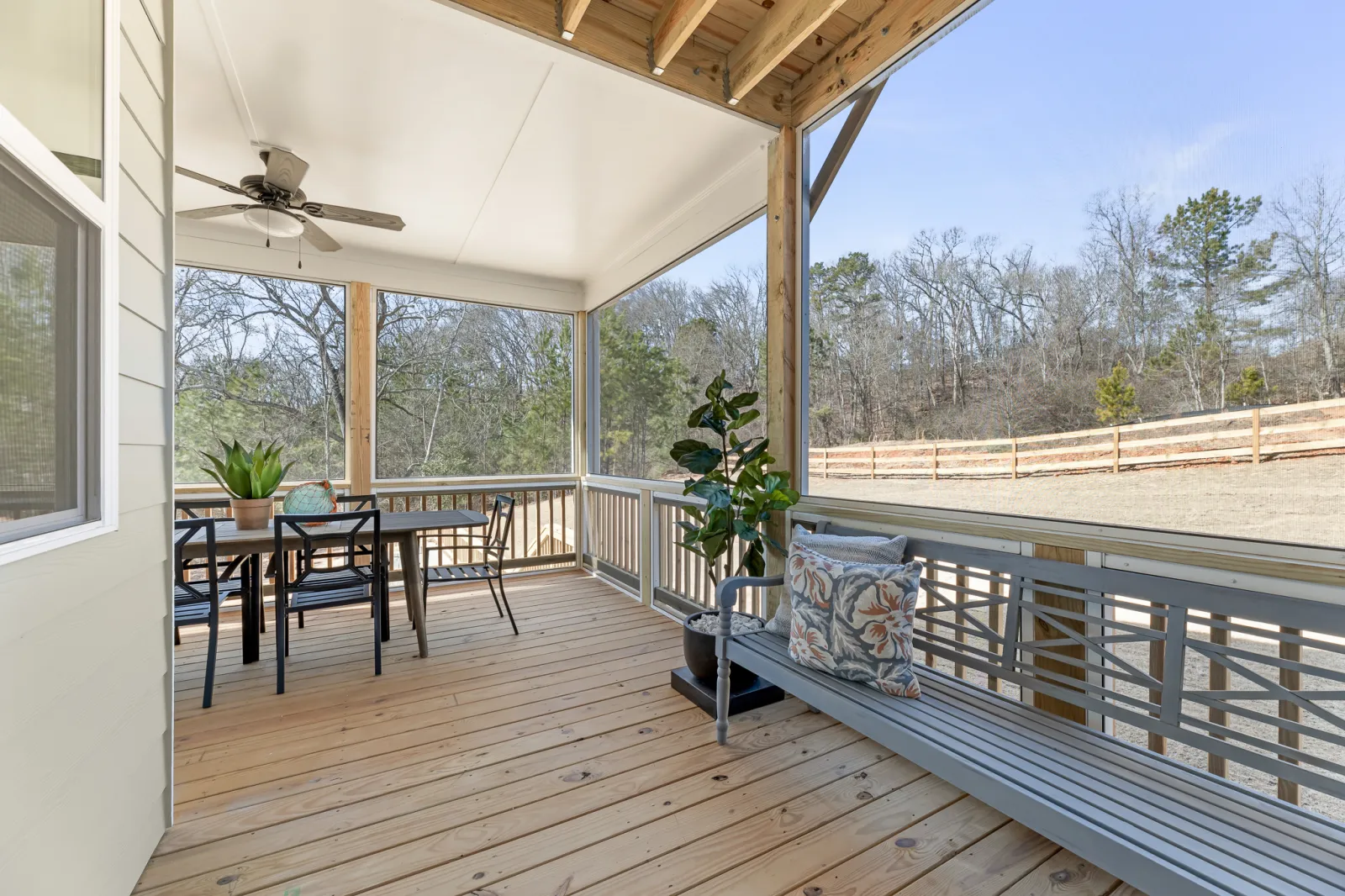 Spacious screened porch with wooden floor, ceiling fan, dining table, bench, plants, and wooded backyard view.