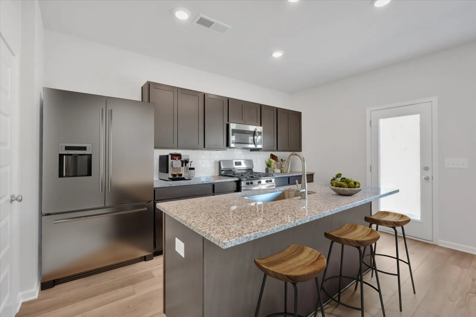 Modern kitchen with granite island, stainless steel appliances, dark cabinets, and three wooden bar stools under bright lighting.