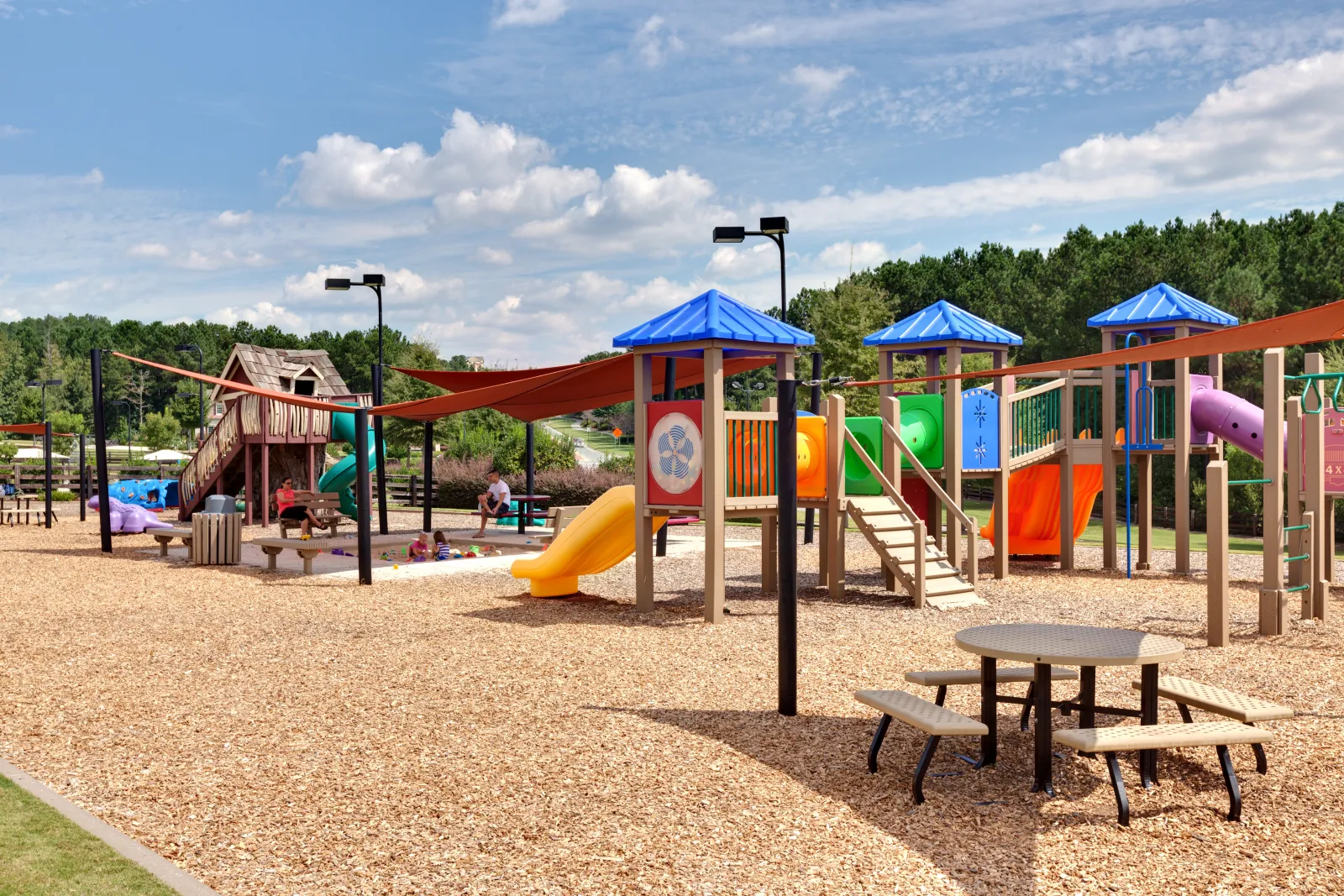 Colorful playground with slides, climbing structures, picnic tables, wood chip ground, and blue sky with clouds.