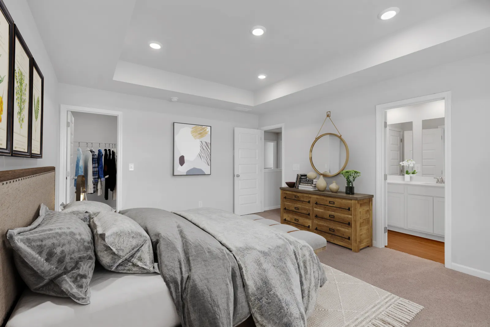 Modern bedroom with gray bedding, wooden dresser, round mirror, and connected bathroom with white vanity.