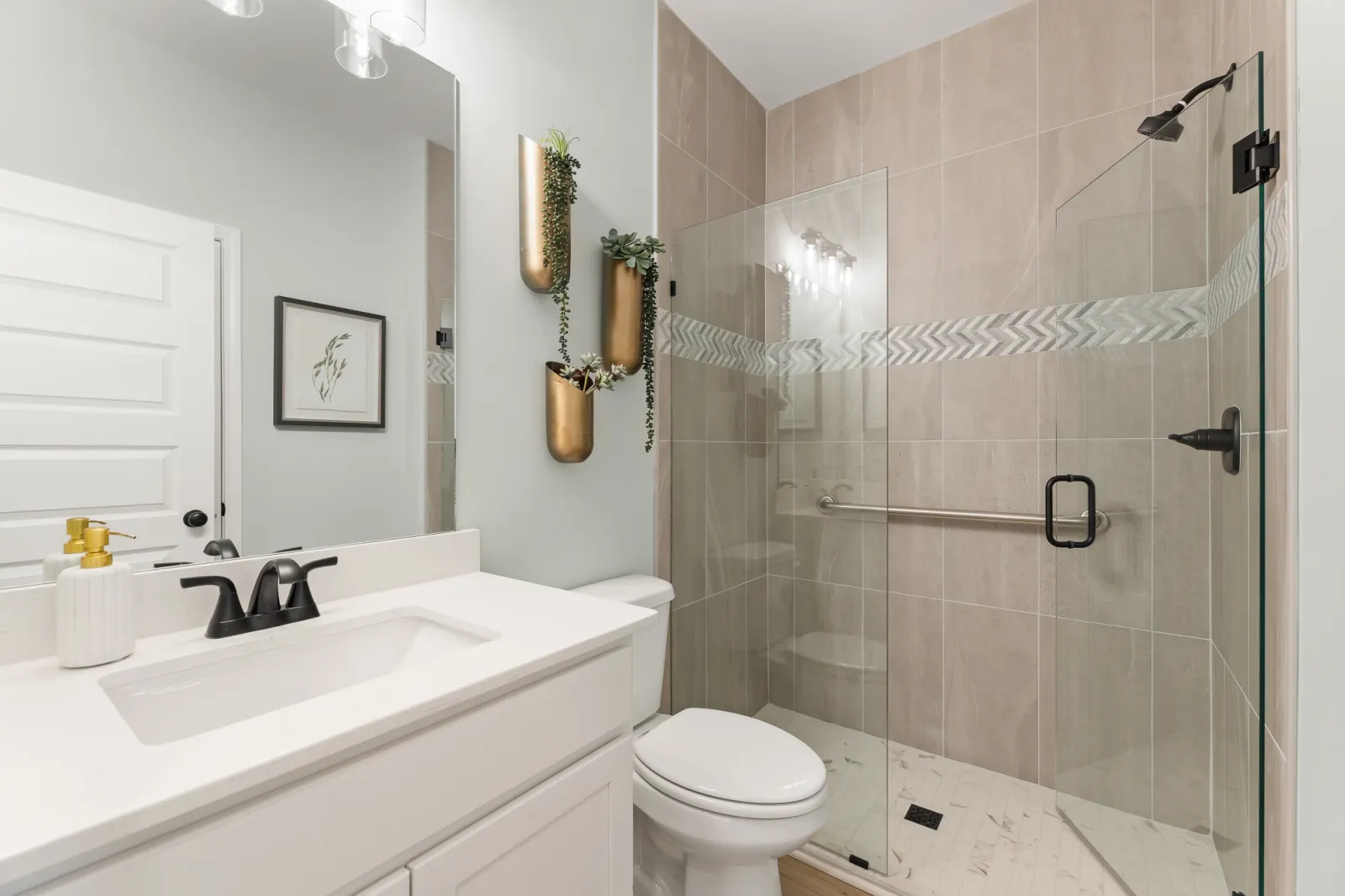 Modern bathroom with glass shower, white vanity, black fixtures, and decorative gold wall planters.