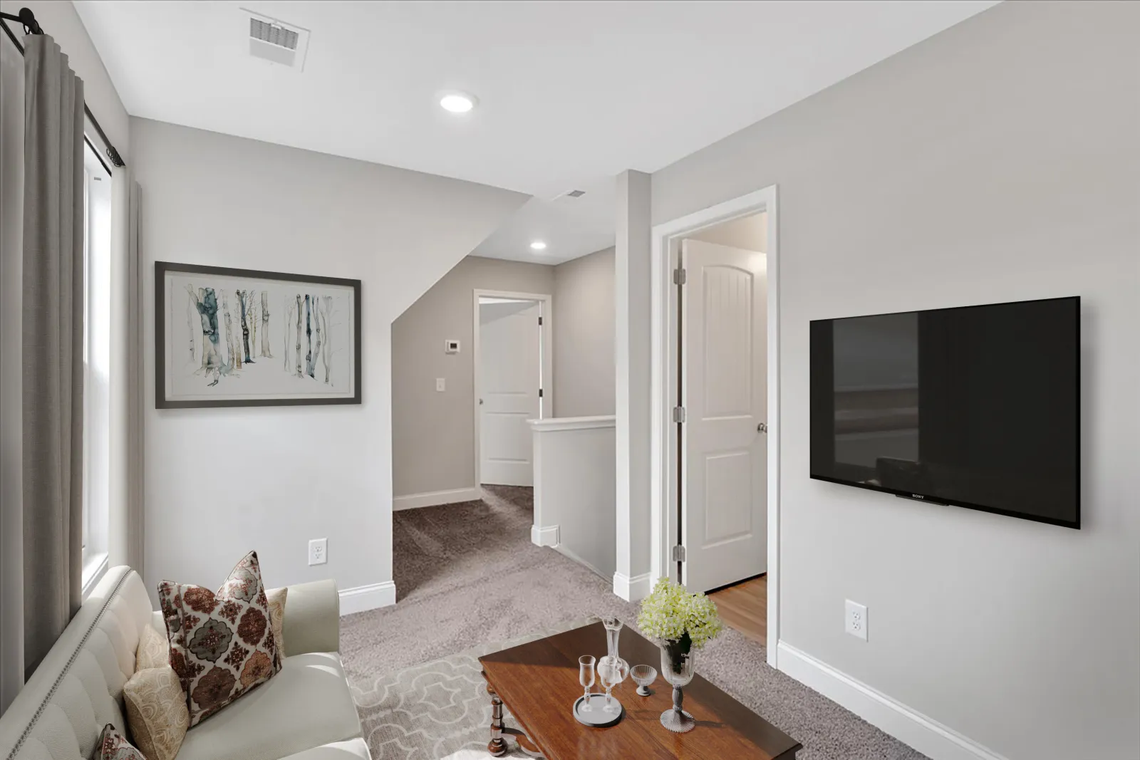 Modern living room with gray carpet, wall-mounted TV, wooden coffee table, light gray walls, and beige couch with pillows.
