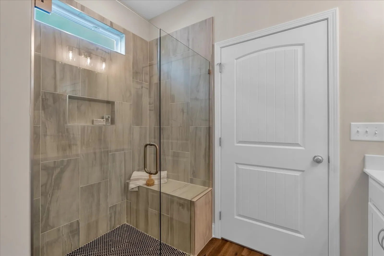 Modern bathroom with glass-enclosed shower, built-in bench, beige tile walls, and white door