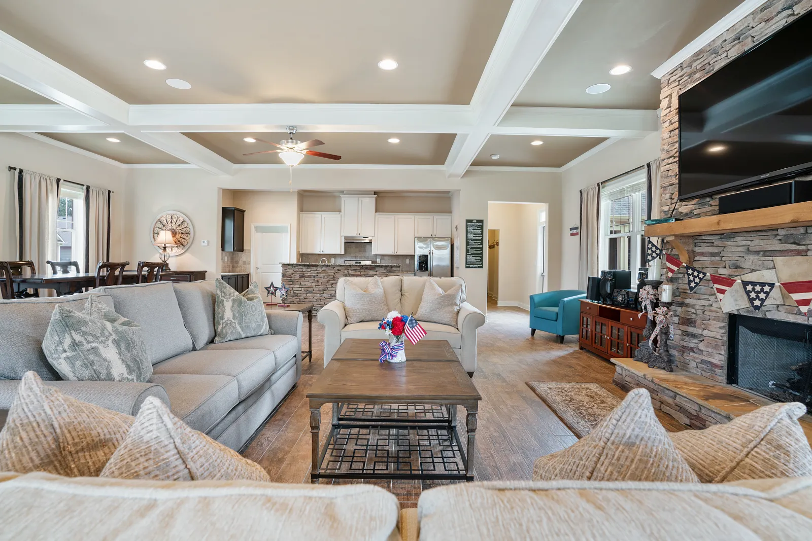 Bright open living room with gray sofas, wooden coffee table, stone fireplace, and kitchen in background.