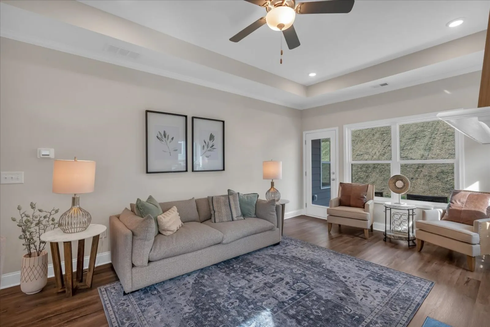 Modern living room with beige sofa, two armchairs, lamps, ceiling fan, and large window with natural light