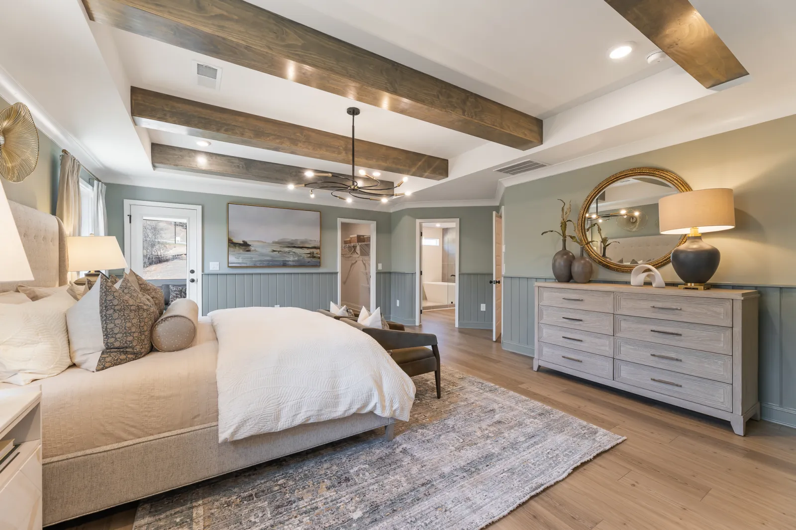 Spacious bedroom with wooden ceiling beams, light bedding, blue-gray walls, and a large dresser with round mirror.