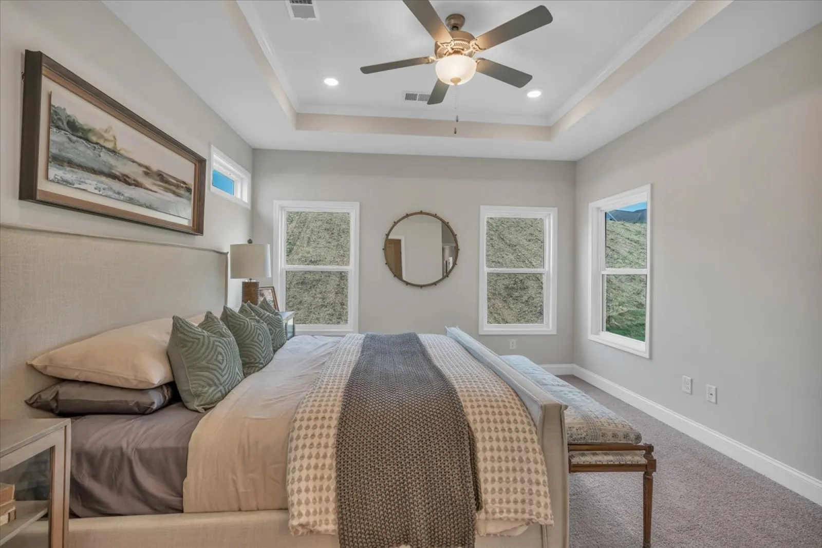 Modern bedroom with beige walls, large bed, patterned blankets, green pillows, ceiling fan, and three windows.