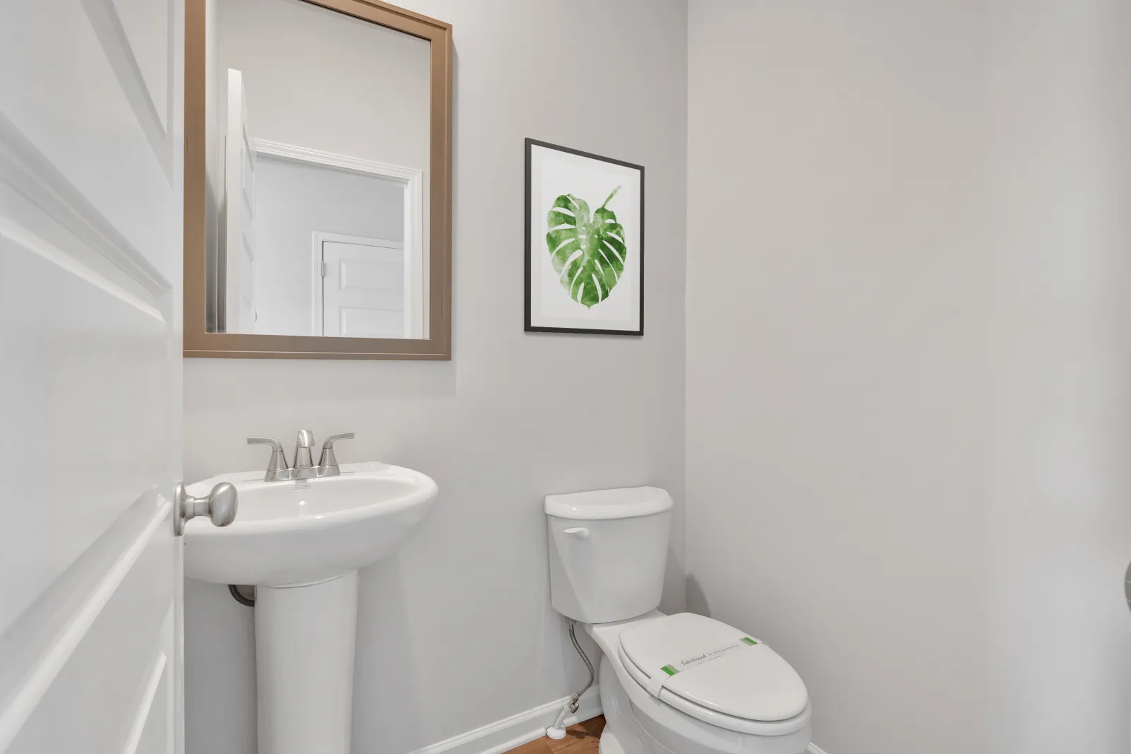 Minimalist bathroom with white pedestal sink, toilet, mirror, and green leaf wall art on gray walls.