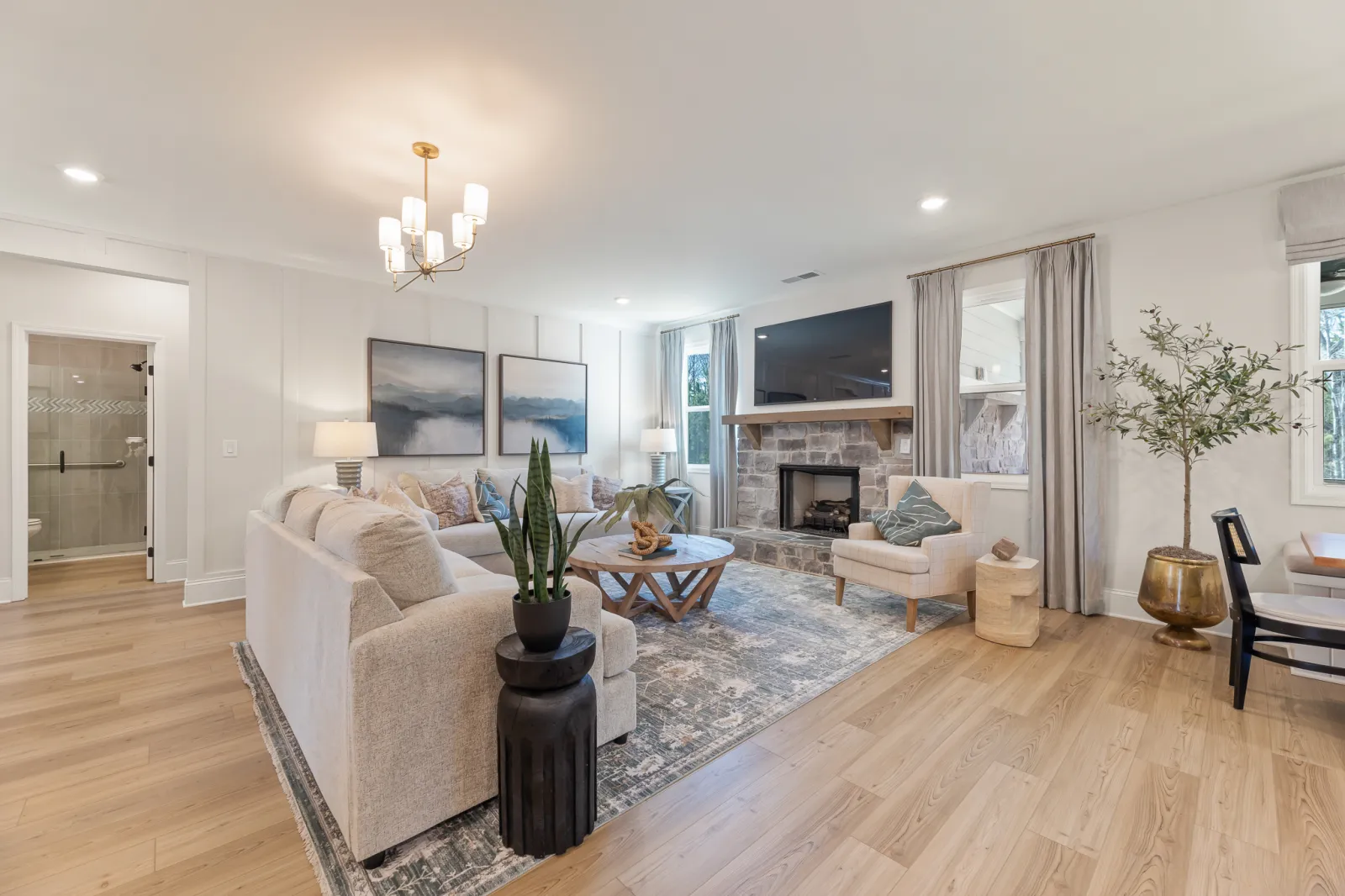 Bright living room with beige sofa, stone fireplace, hardwood floors, modern chandelier, and large windows with curtains.