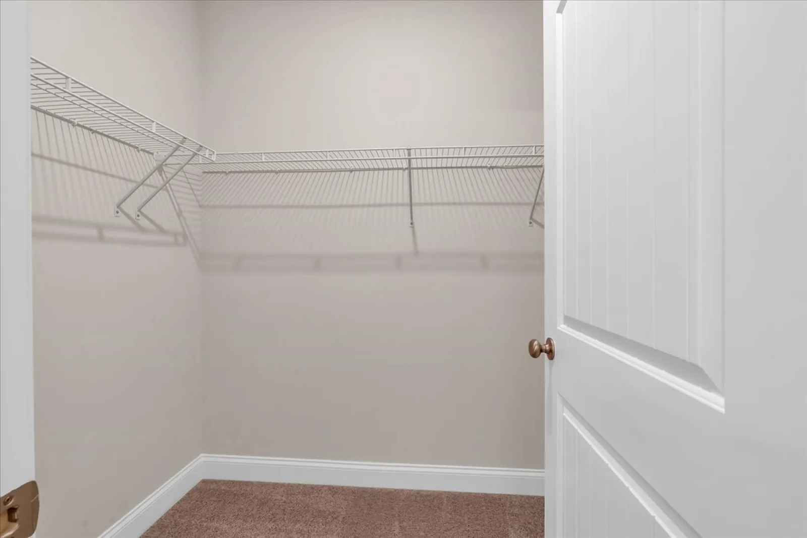 Empty walk-in closet with beige walls, carpeted floor, white wire shelves, and a partially open white door.