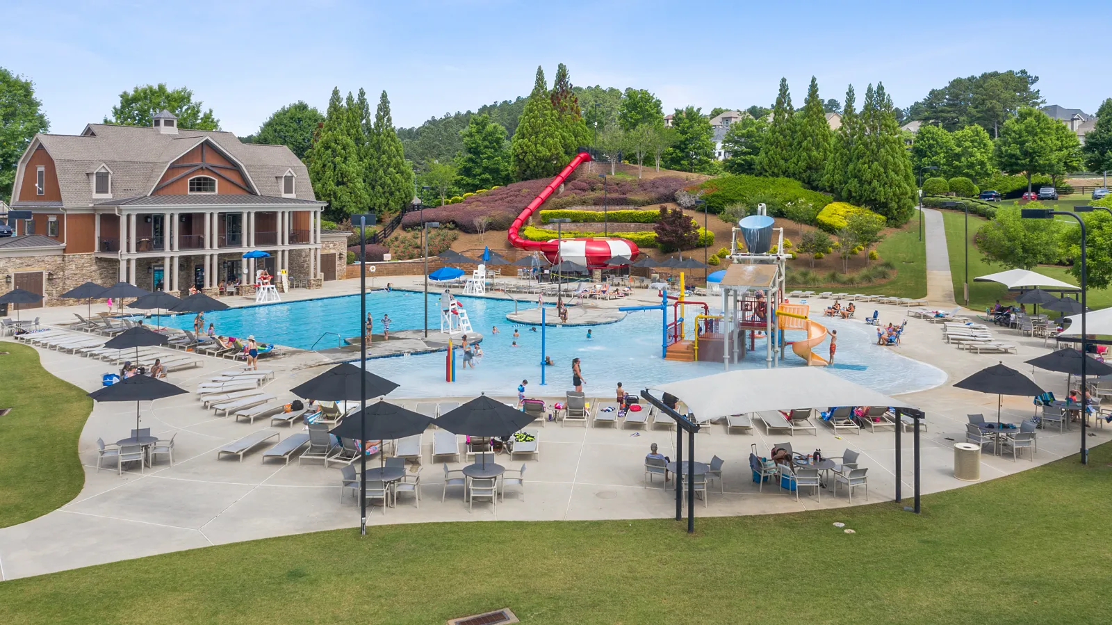 Outdoor community pool with water slides, splash pads, lounge chairs, umbrellas, and a clubhouse on a sunny day.