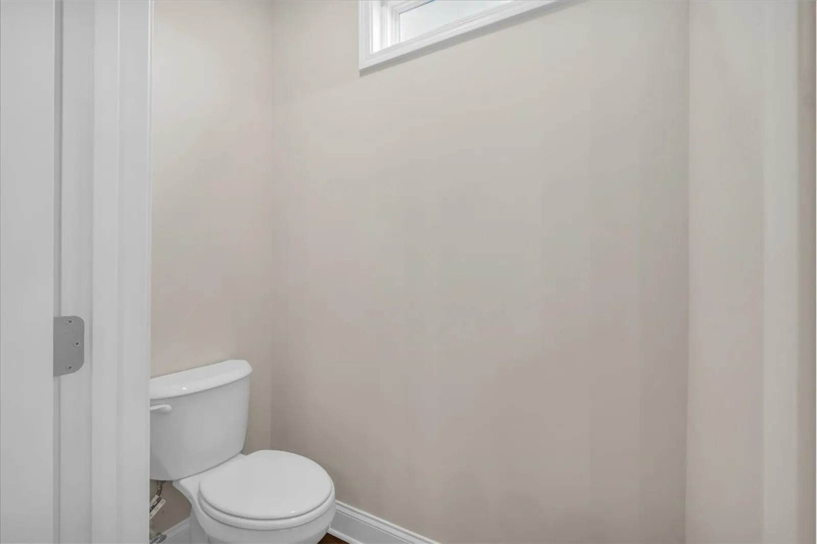 Small clean bathroom with white toilet and beige walls under a small high window.