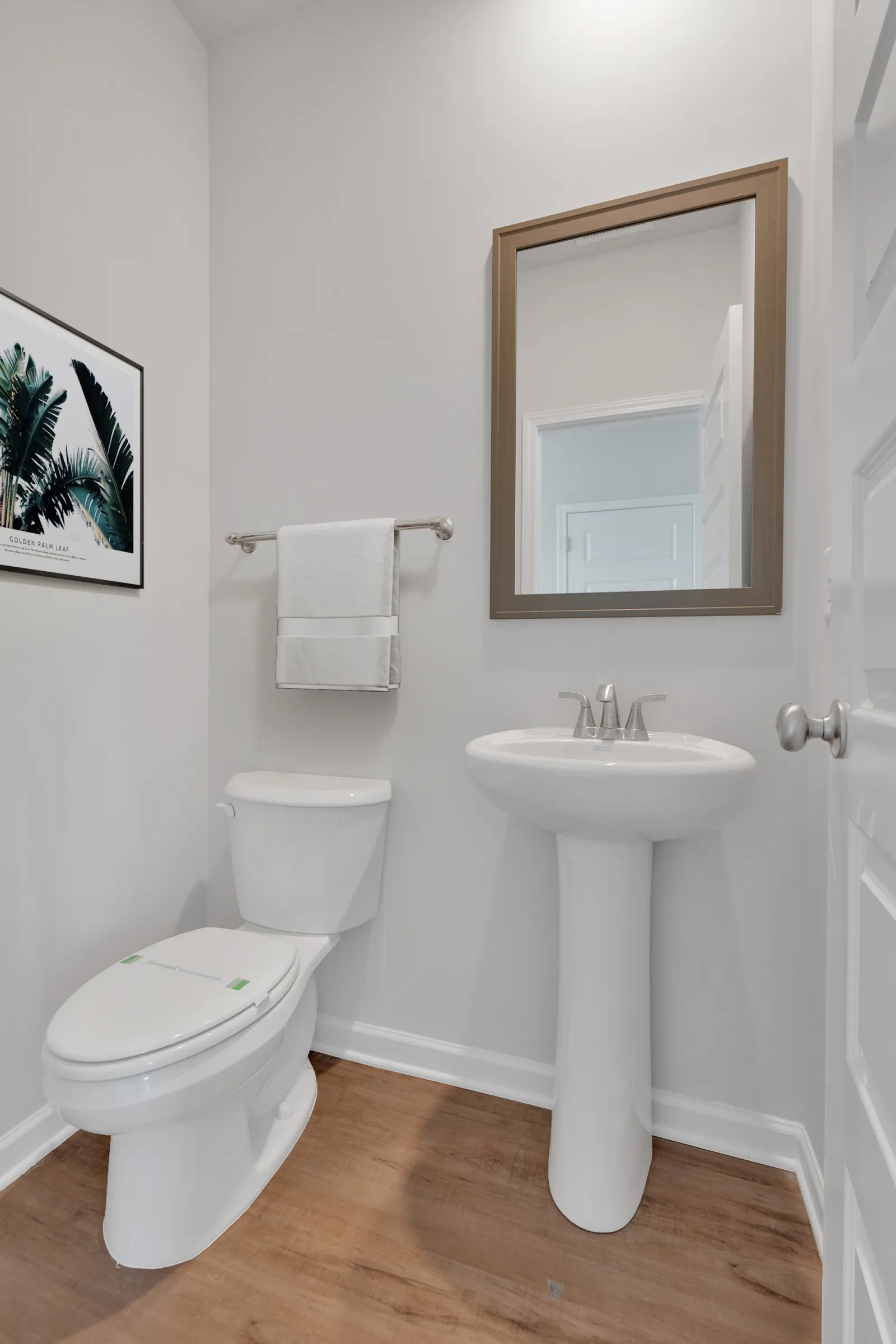 Bright small bathroom with white toilet, pedestal sink, large mirror, towel rack, and wall art on wood floor.