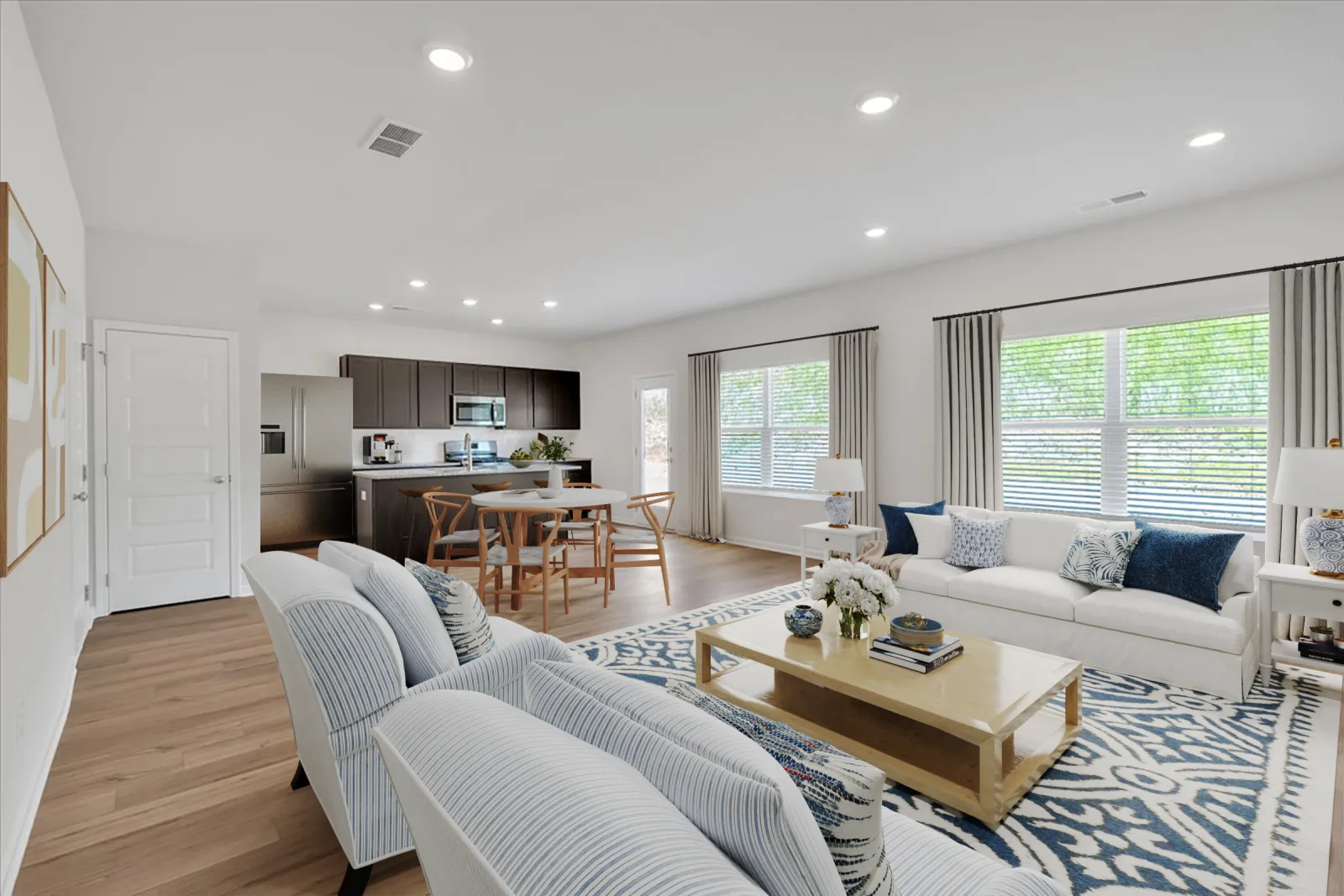 Bright open-concept living room and kitchen with white sofas, wooden dining table, and large windows with curtains.
