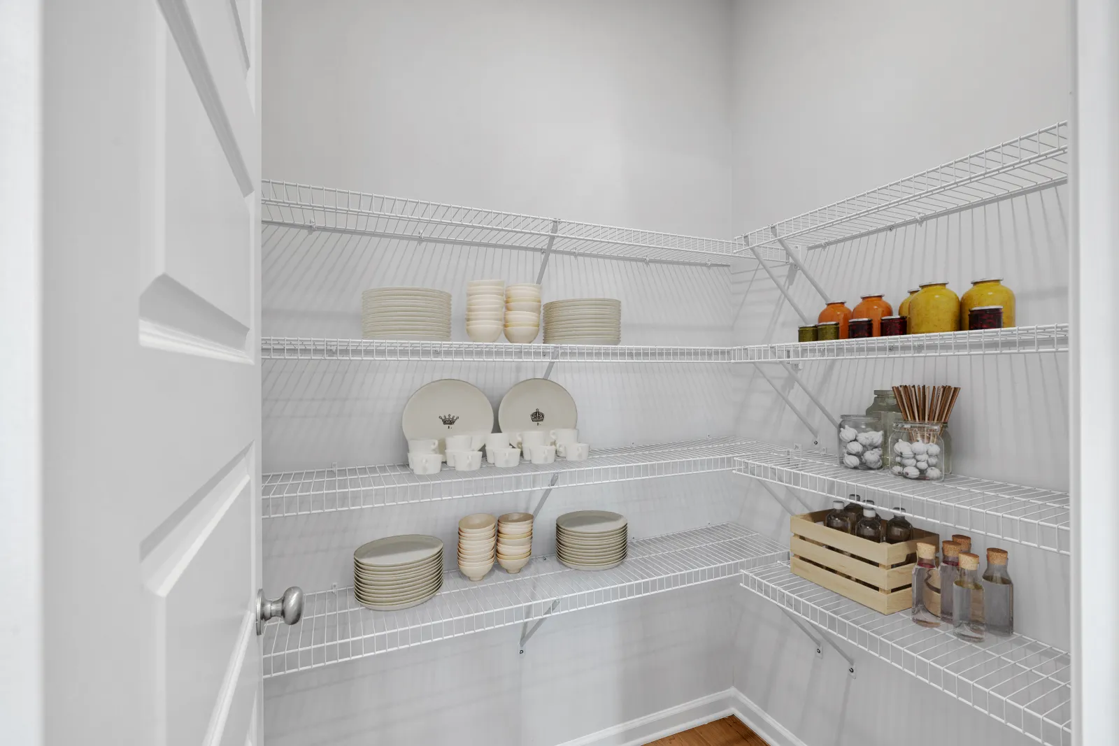 Organized pantry with white wire racks holding plates, mugs, jars, and bottles in a clean white room.