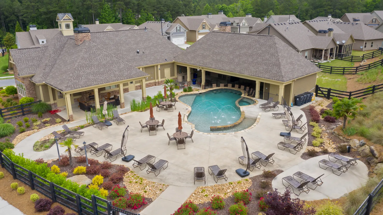 Community pool area with loungers, umbrellas, tables, and clubhouse surrounded by houses and landscaping