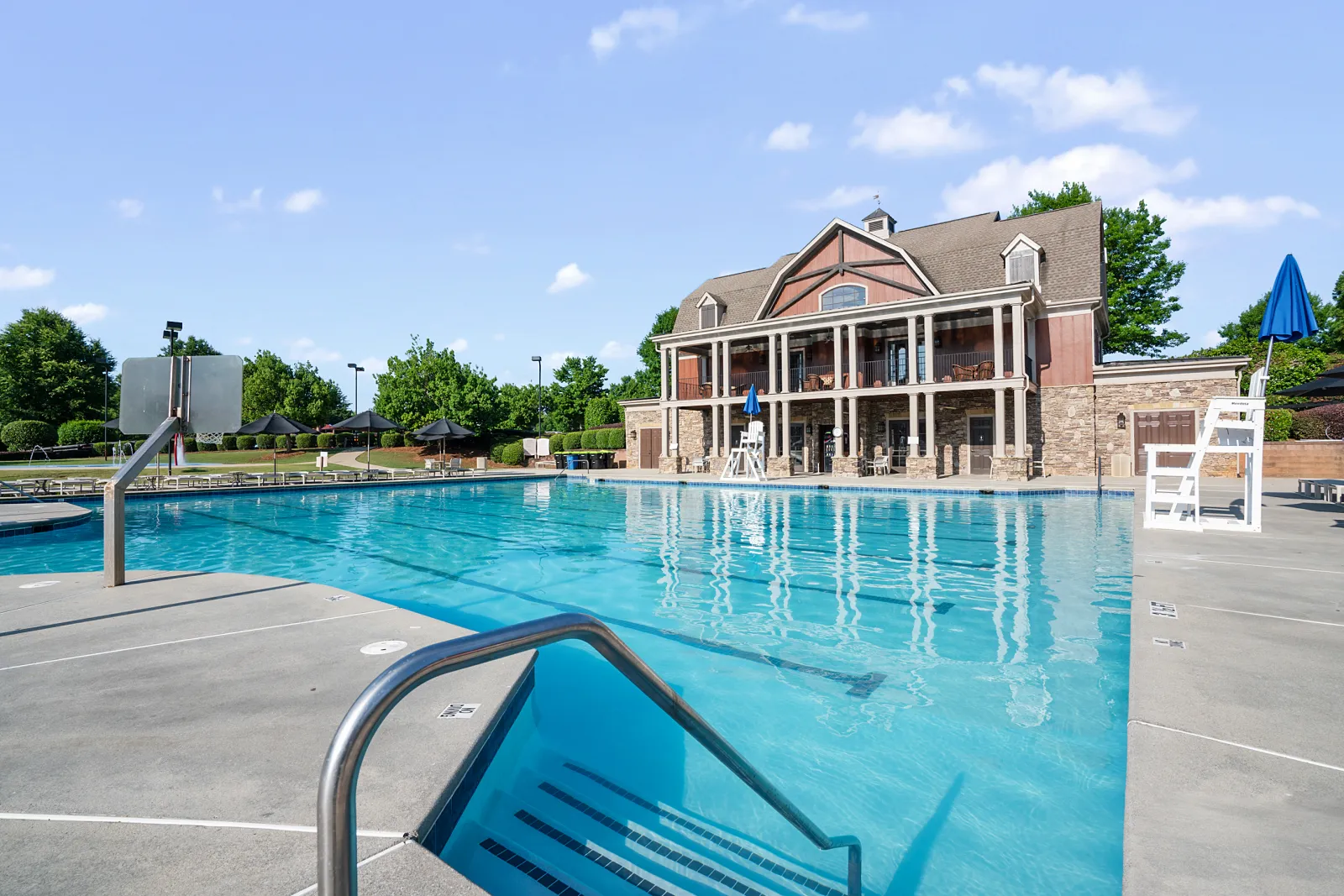 Outdoor swimming pool with clear blue water, poolside chairs, and a large clubhouse on a sunny day.