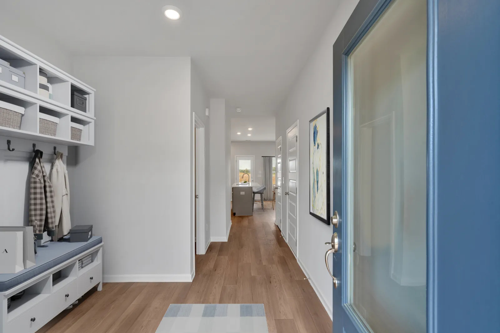 View of a modern entryway with a blue door, white walls, wooden floor, and hallway leading to a kitchen area.